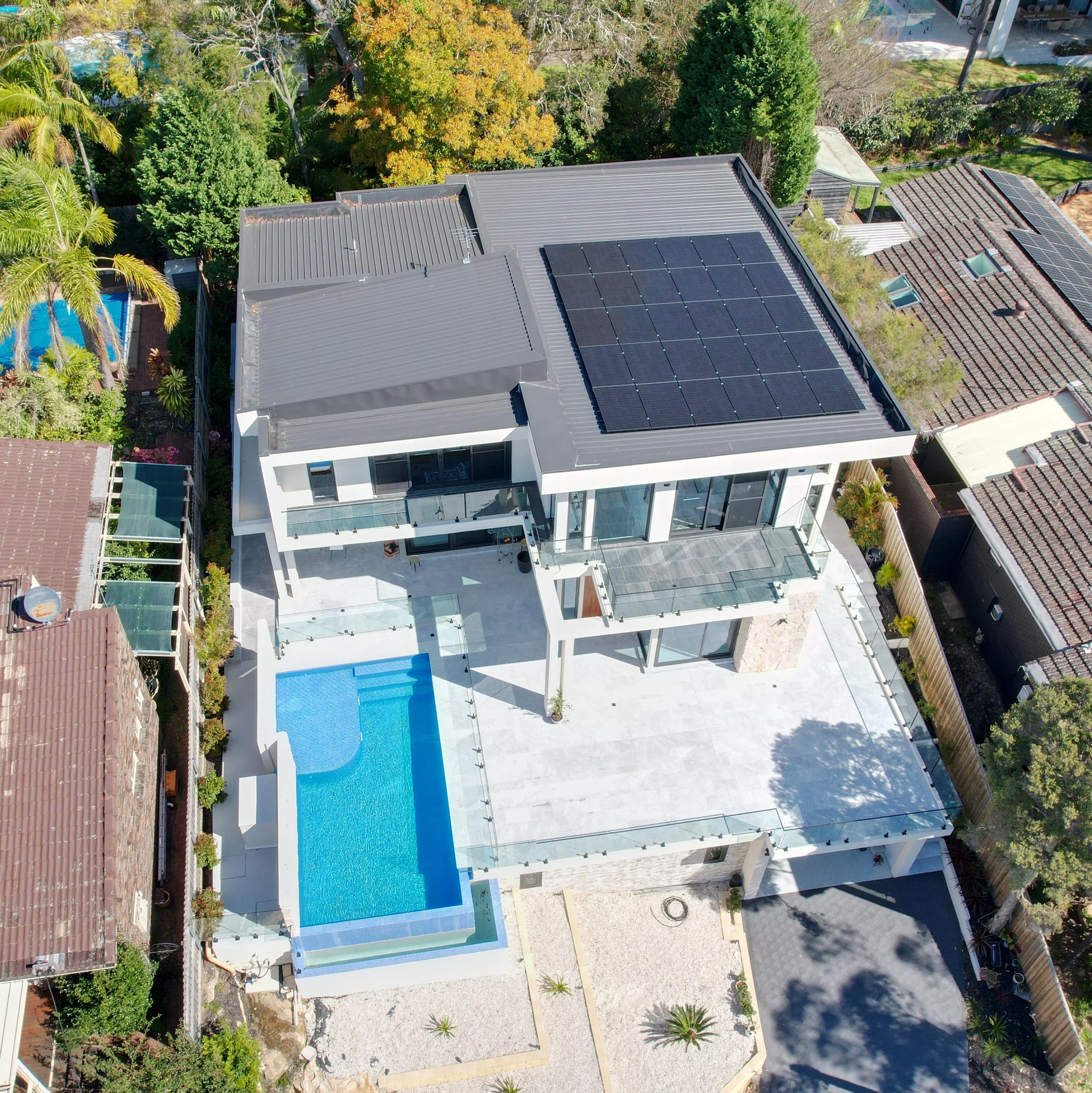 Aerial view of a modern house with rooftop solar panels, a swimming pool, and a spacious backyard, surrounded by neighboring houses and lush greenery.