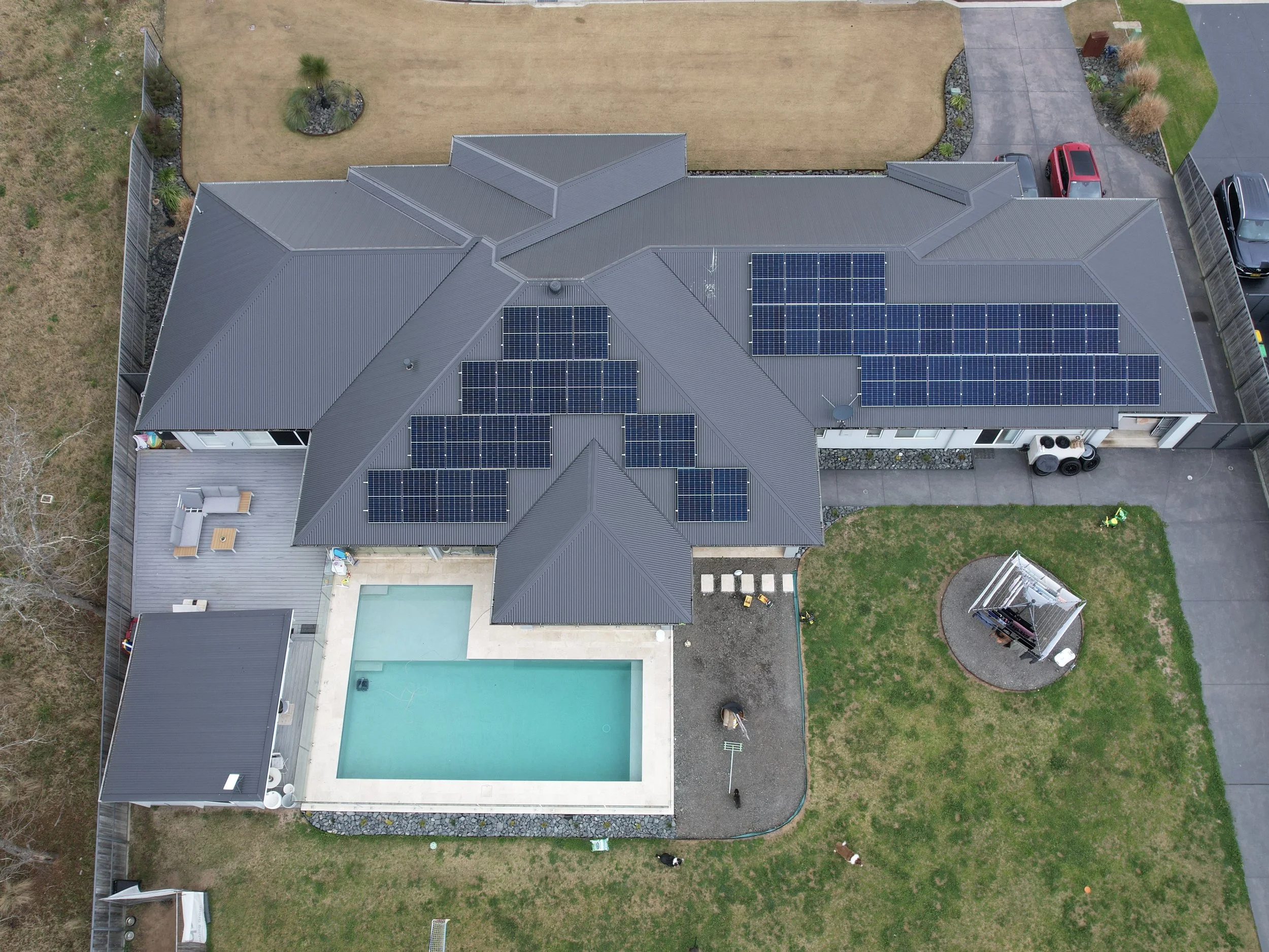 Aerial view of a house with a gray roof, solar panels, a backyard with a swimming pool, a deck with outdoor furniture, and a small circular gazebo with a white canopy.
