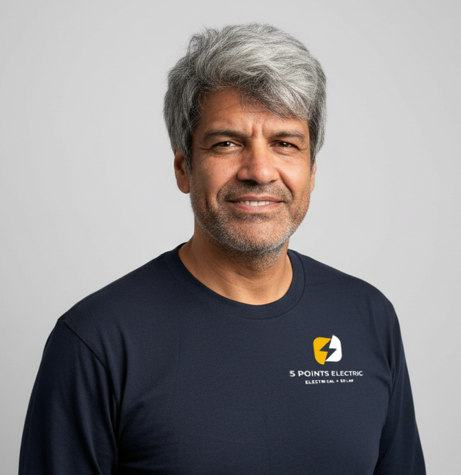 A middle-aged man with gray hair and a beard, smiling, wearing a navy blue t-shirt with a logo and text that reads '5 POINTS ELECTRIC' against a plain light gray background.