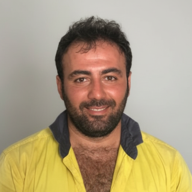A man with dark hair, a beard, and moustache, smiling, wearing a yellow shirt with a dark collar, standing against a plain light-colored wall.