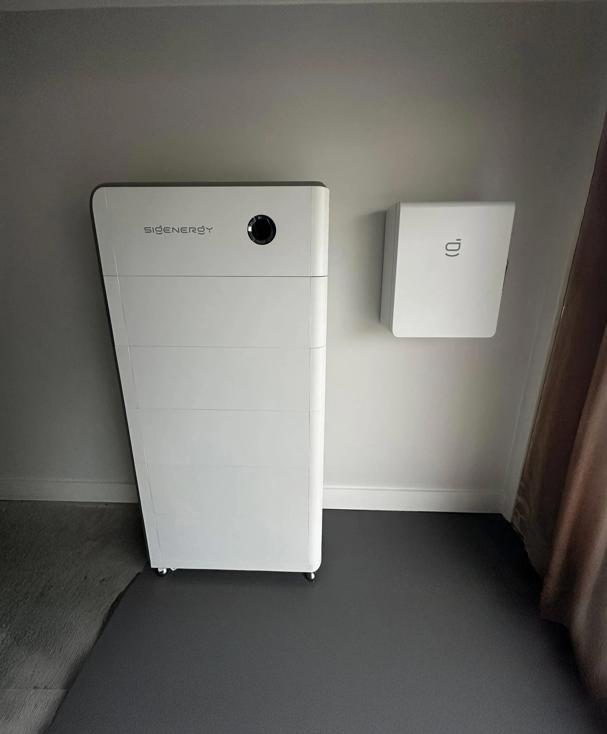 A white, modern air purifier or dehumidifier with the brand name 'sigenergy' on it, placed on small wheels, next to a wall with a white box mounted on it, near a brown curtain and gray carpet.