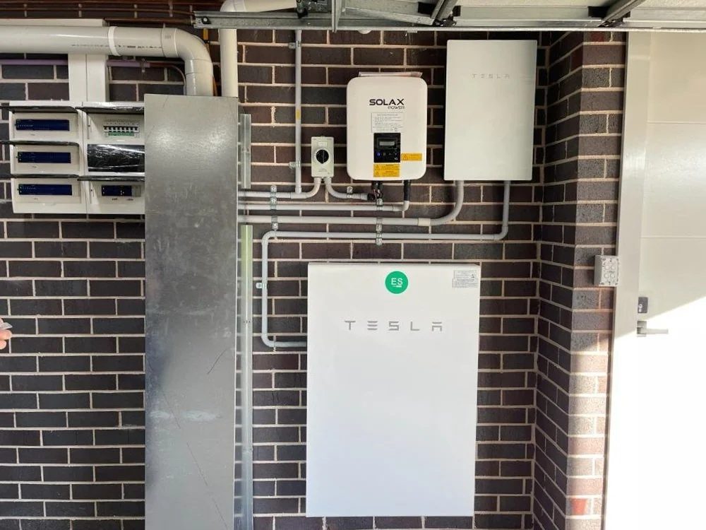 Electrical and solar equipment mounted on a brick wall, including Tesla inverter, solar power controller, and wiring panel.