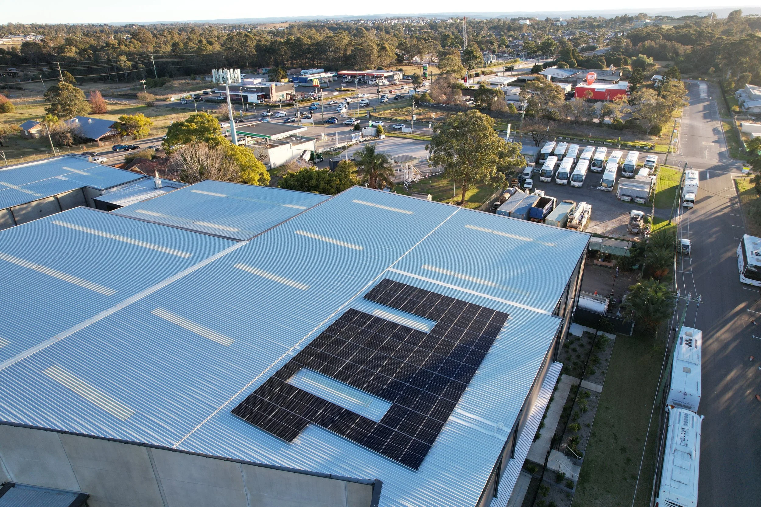 Aerial view of a commercial building with solar panels on the roof, surrounded by parking lots, roads, and trees in a suburban area.