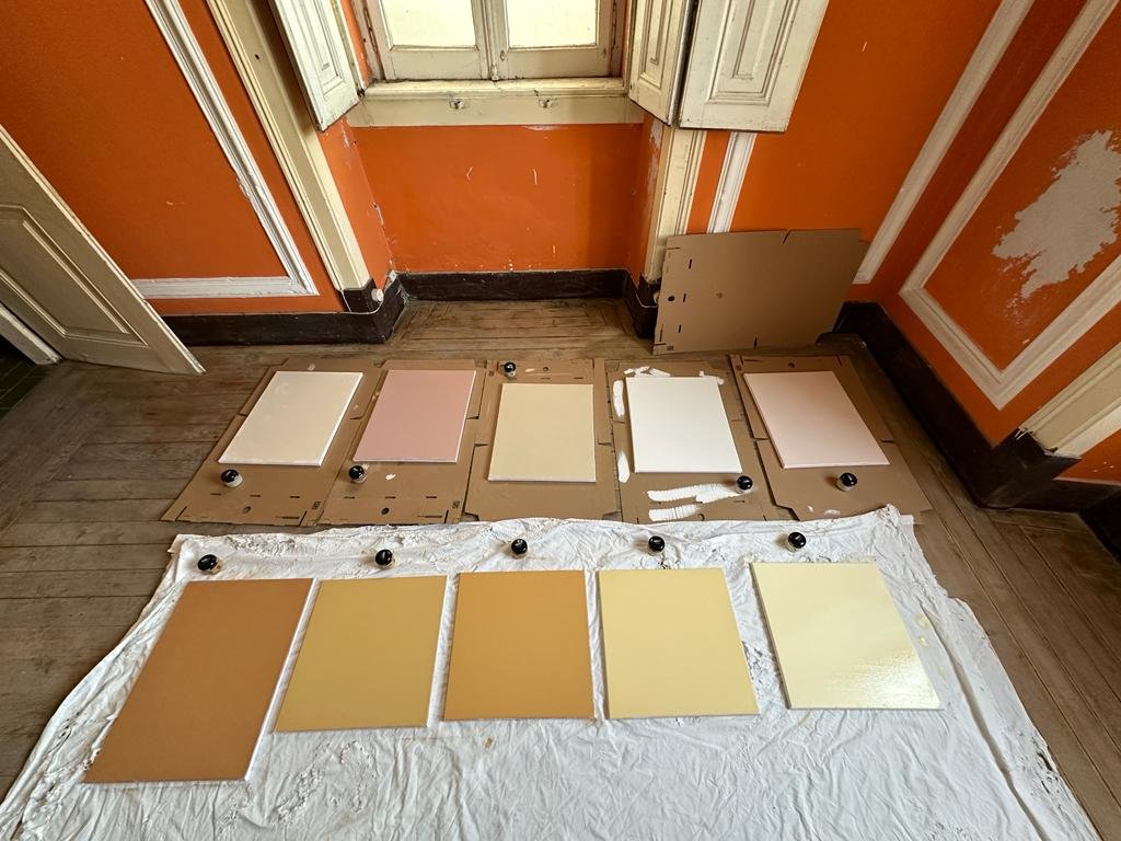 Nine paint samples on canvases are spread out on a drop cloth and cardboard in an orange-walled room with wooden floors.