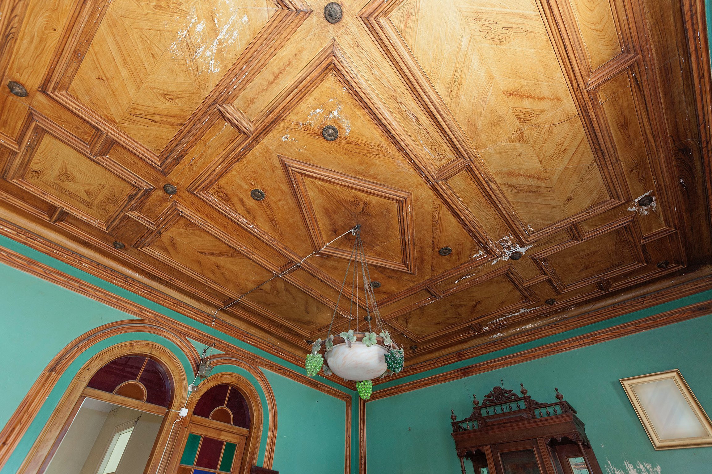 Ornate wooden ceiling with decorative molding in a room with teal walls, a vintage chandelier, arched doorway, and antique furniture.