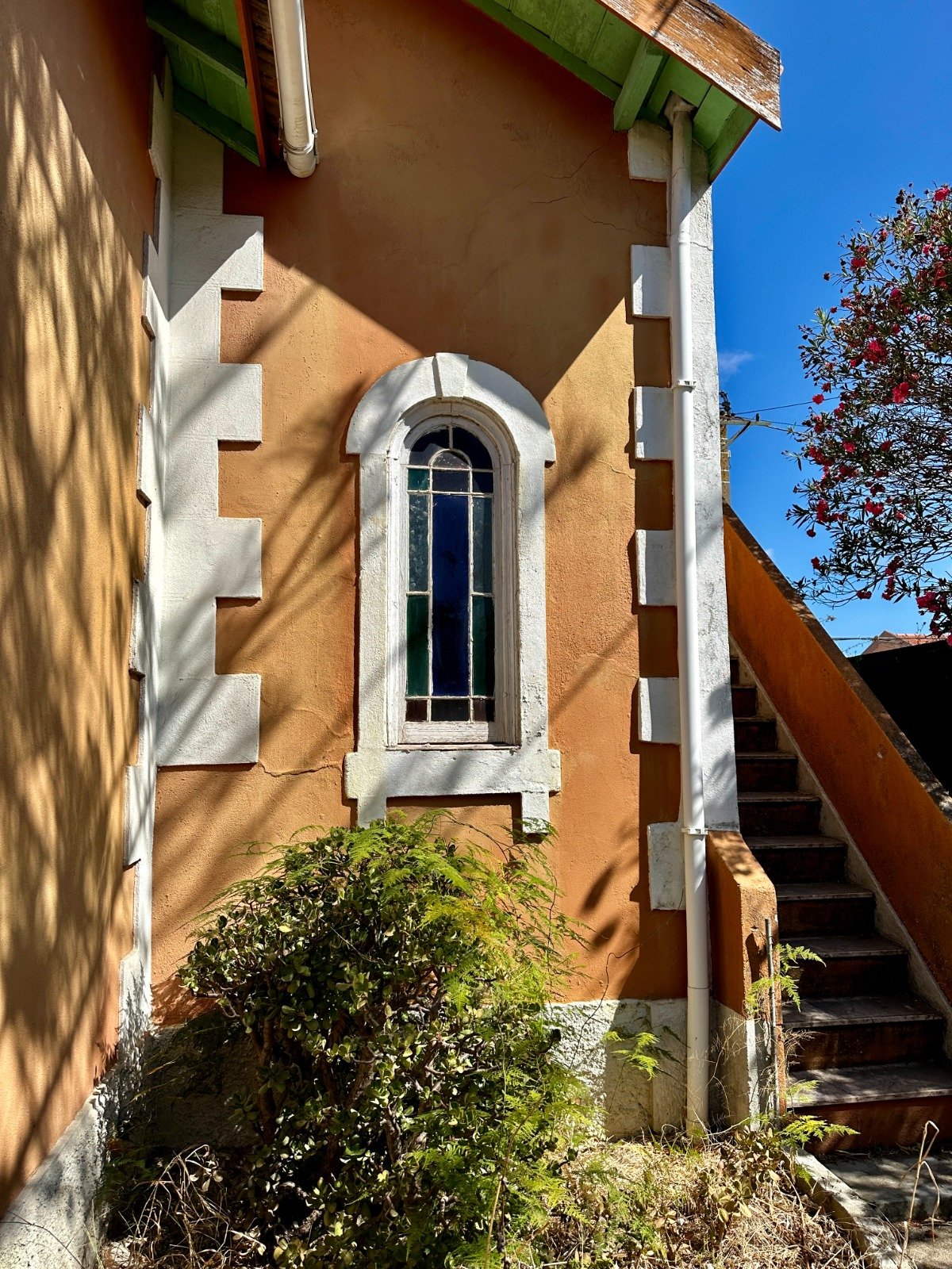 Vintage-style building with arched window, orange facade, white trim, exterior staircase, and greenery, under blue sky