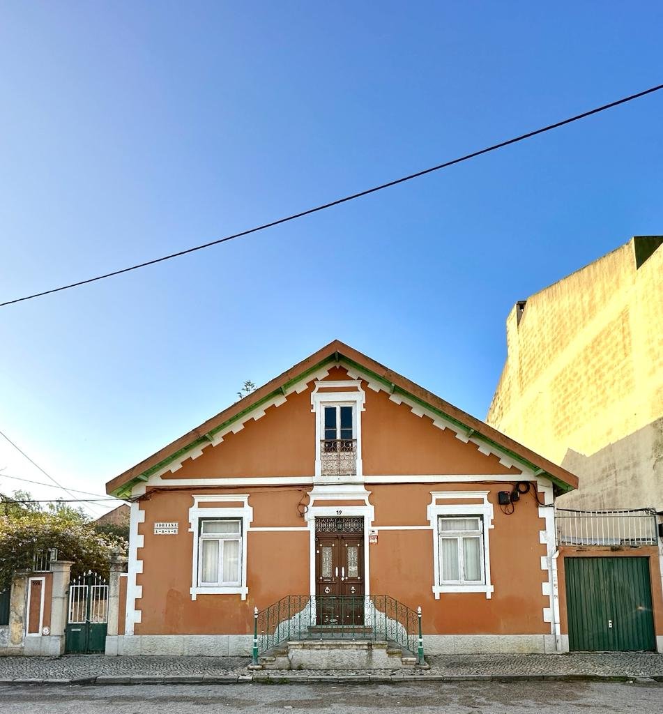 Historic orange house with white trim, wooden door, and two windows, set against a clear blue sky. A gate is adjacent to the house.