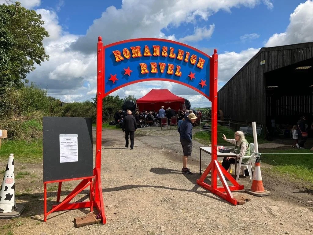 Entrance sign for Romansleigh Revel with a brass band gathered under a red tent, some seated, some standing, on a farm with a barn, trees, and cloudy sky in the background.