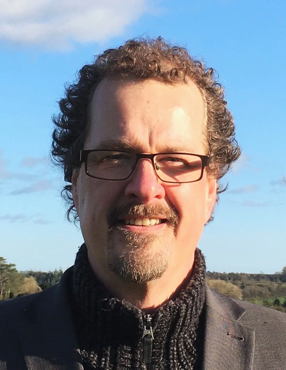 A man with curly hair, glasses, and a goatee smiling outdoors on a sunny day with blue sky and some clouds in the background.