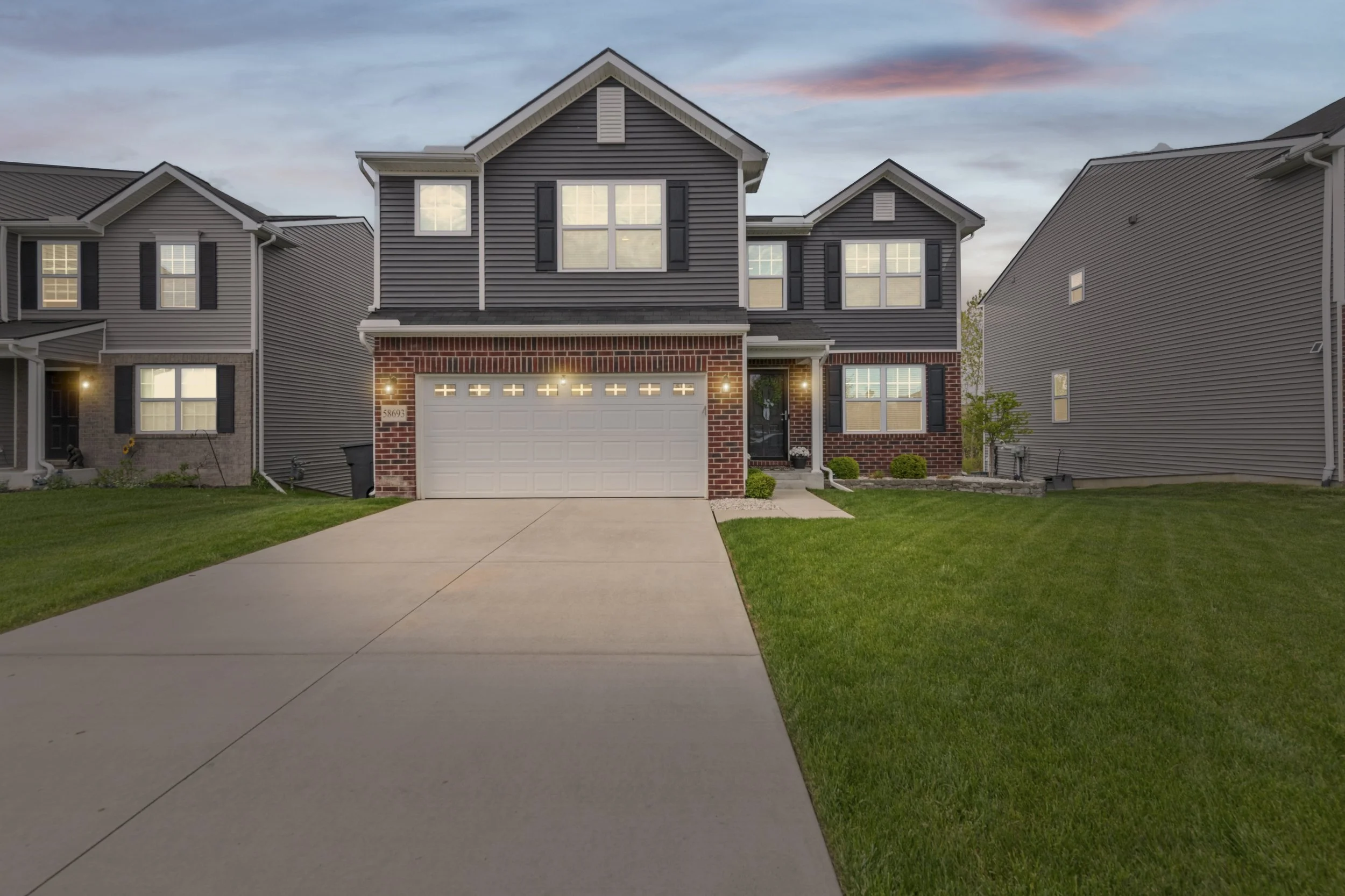 Front view of a modern two-story house with a brick and horizontal siding exterior, a white garage door, black shutters, and a small front porch, with neighboring houses on each side and a well-maintained lawn, under a sky with pink and blue hues dur