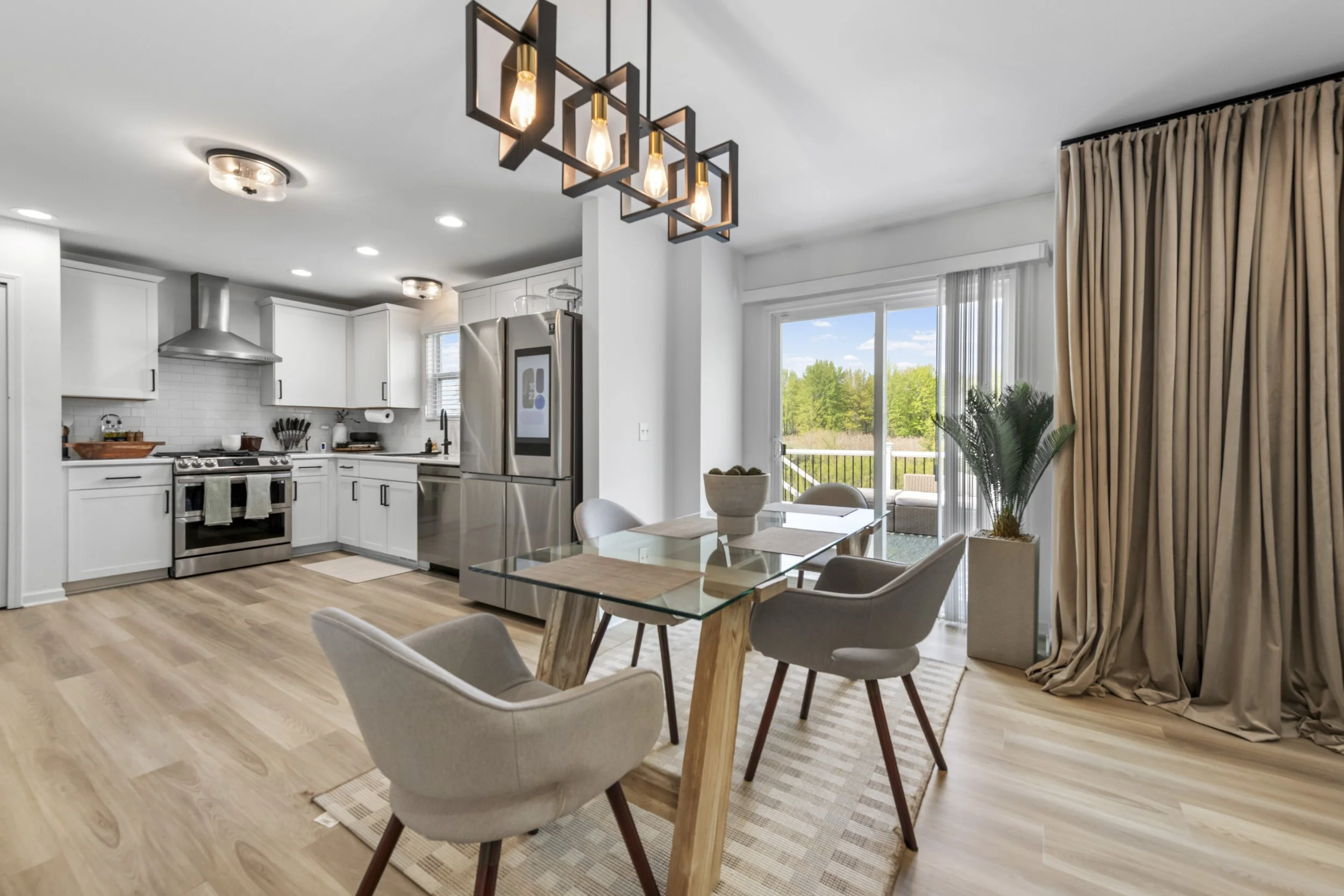 Modern kitchen and dining area with white cabinets, stainless steel appliances, light wood flooring, and large sliding glass door with beige curtains leading to outdoor balcony with green trees.