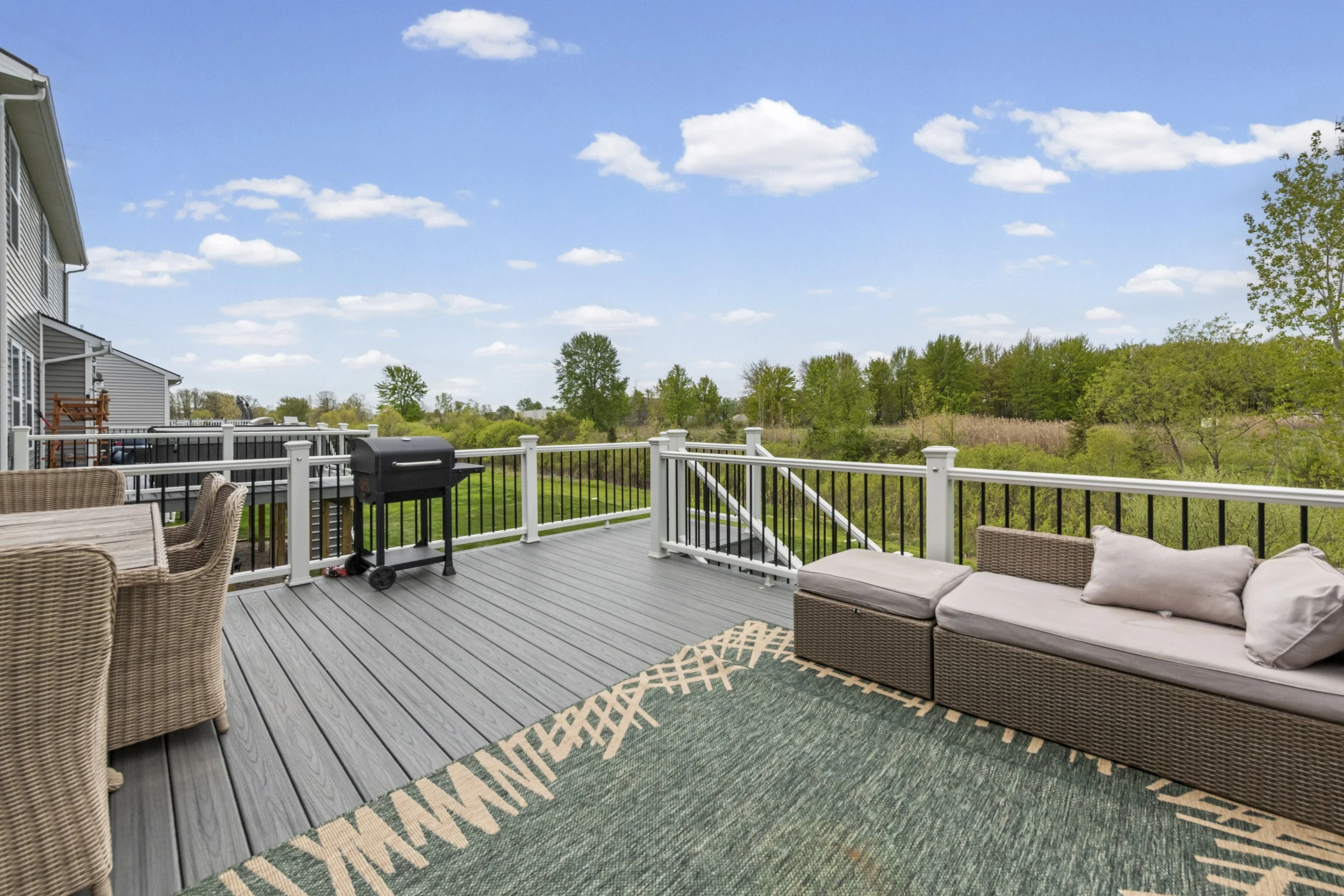 A spacious outdoor deck attached to a house, featuring wicker chairs, a lounge sofa with cushions, a green and beige outdoor rug, a black grill, and a safety railing, overlooking a lush green landscape and a bright blue sky with scattered white cloud