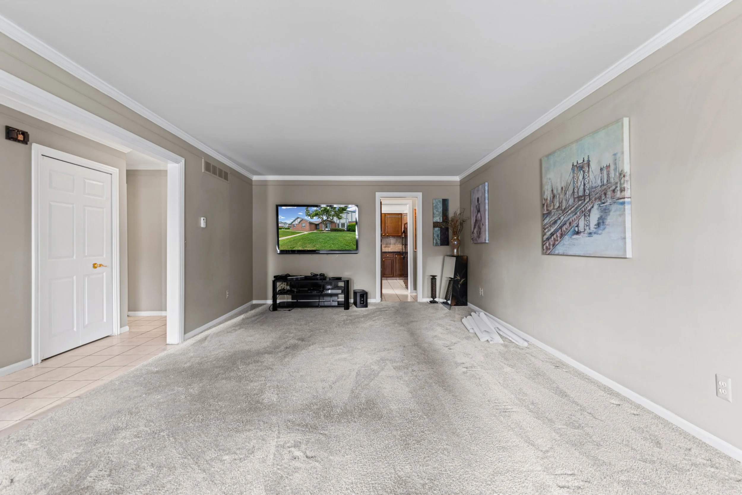 Empty living room with beige carpet, light gray walls, and white crown molding. There are paintings on the wall, a small black TV stand with a television, and a doorway leading to a kitchen area.