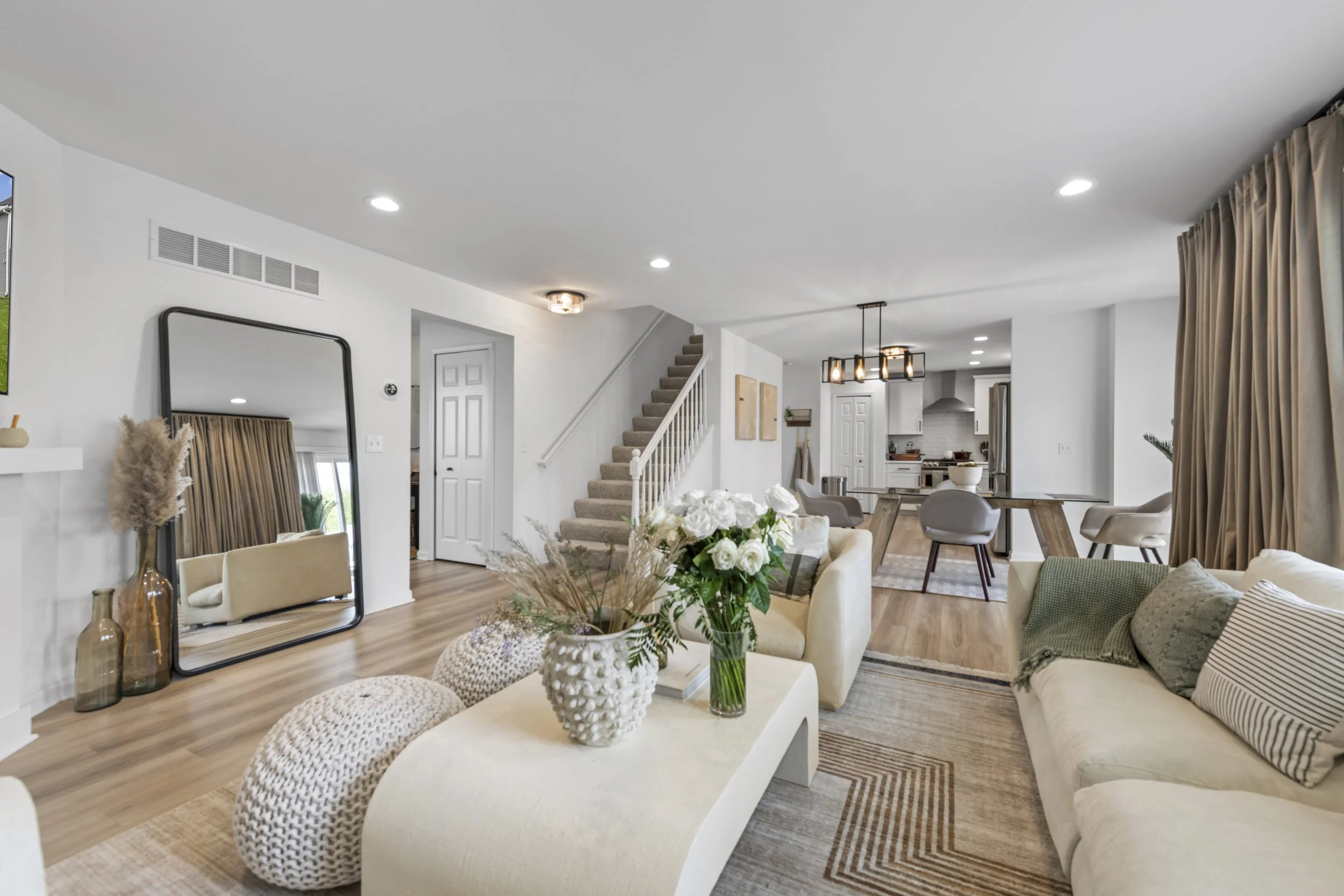Open-concept living room and kitchen with staircase, decorated in neutral tones, featuring a large mirror, floral arrangements, seating area, and pendant lighting.