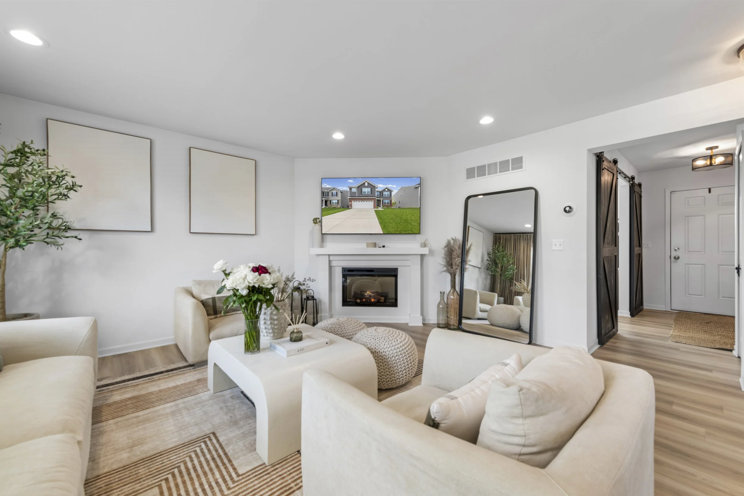 Living room with beige sofas, a white coffee table with flowers, wall art, a fireplace, a large mirror, and a TV showing a house exterior.