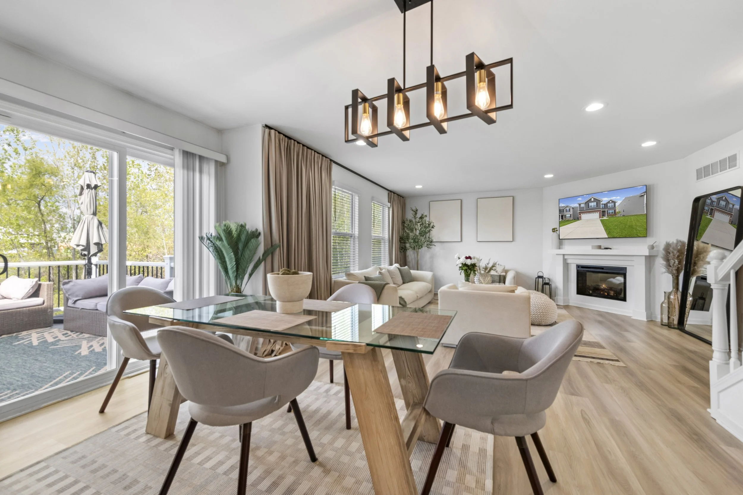 Open-plan living and dining area with large windows, beige curtains, a modern chandelier, a fireplace, and a TV overlooking a backyard with a house and lawn.