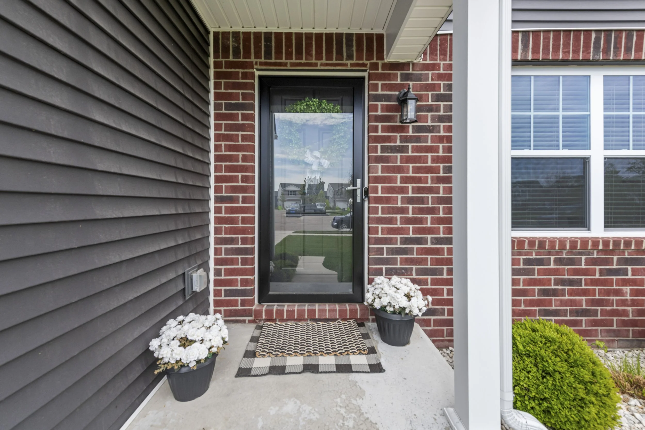 Front porch of a house with a brick wall entry door, potted white flowers, a black doormat, and a black lantern light fixture.