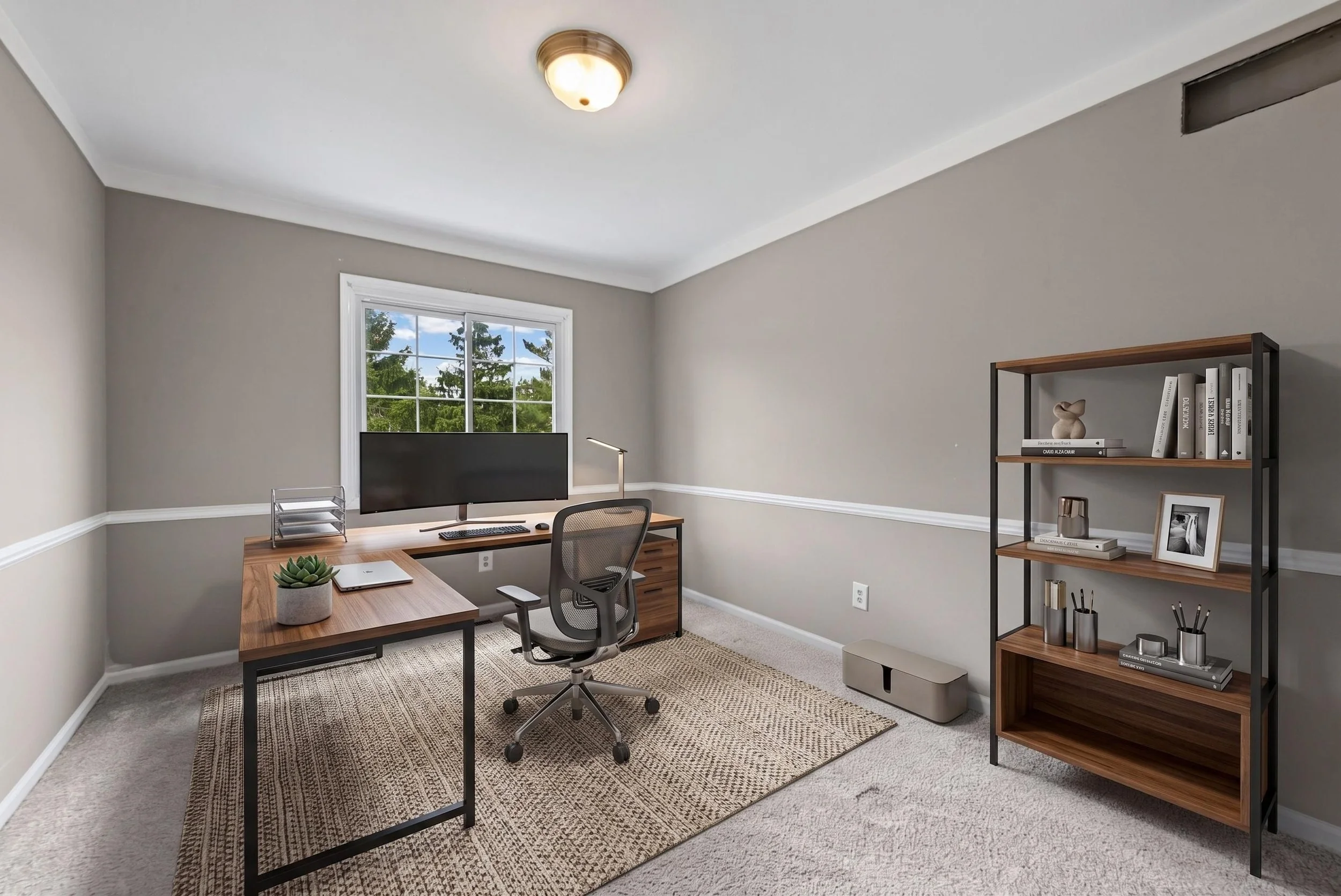 A modern home office with a wooden desk, computer monitor, office chair, bookshelf with books and decorative items, window showing trees outside, ceiling light, and a beige rug on carpeted floor.