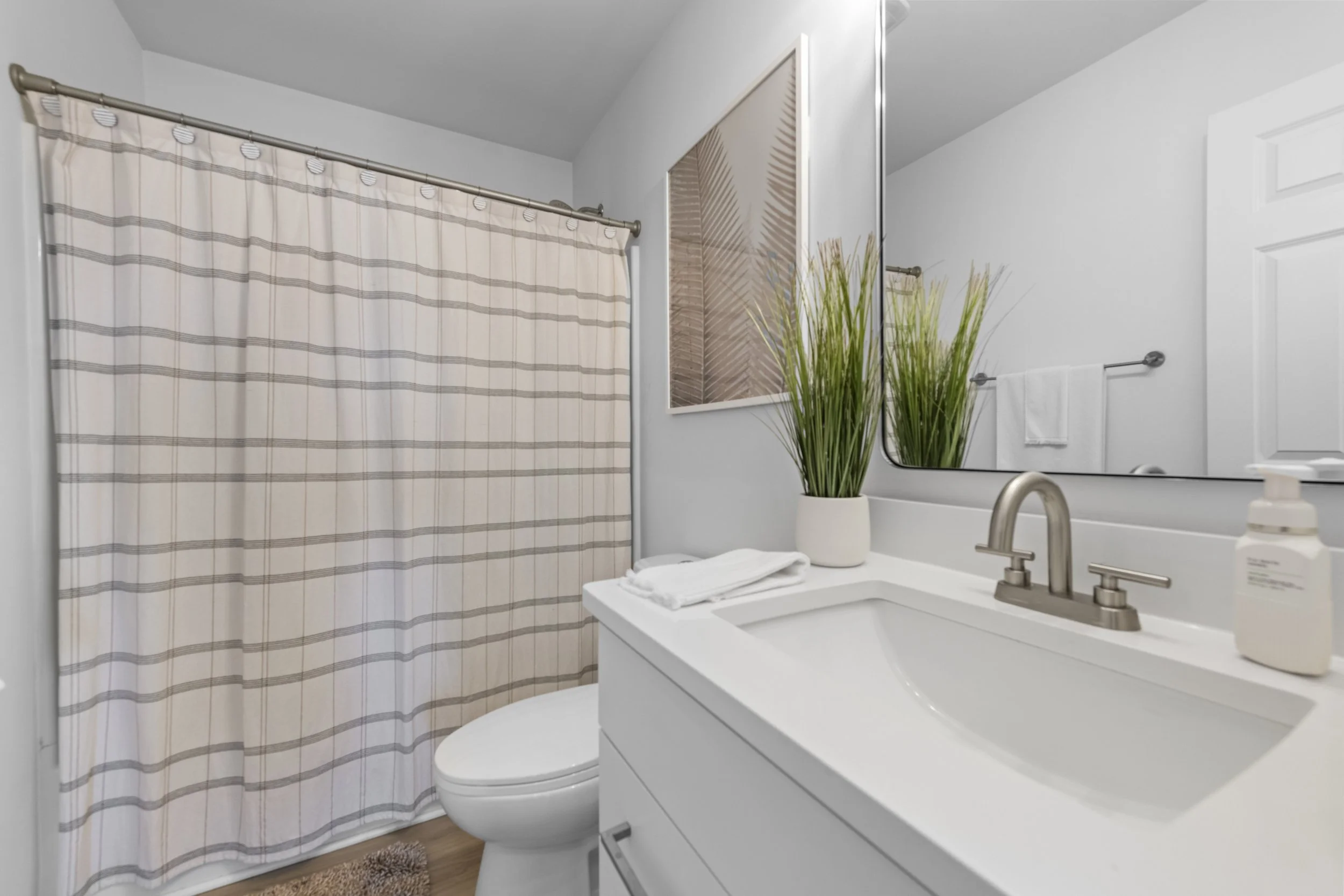 Modern bathroom with a white vanity, a large mirror, a potted plant, a towel, a soap dispenser, and a striped shower curtain.