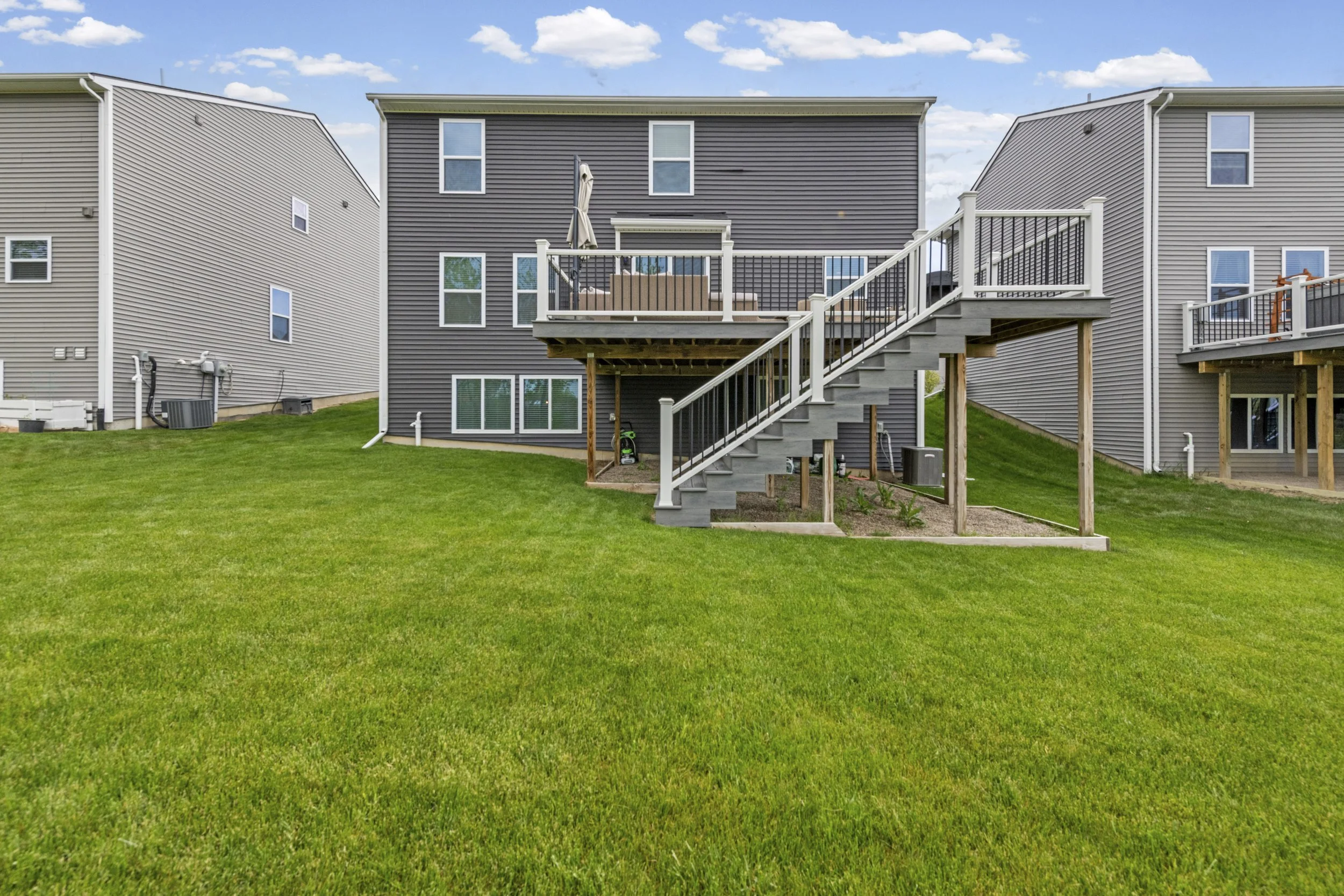 Backyard view of three houses with gray siding, a wooden staircase, a new deck with outdoor furniture, and a lawn with green grass.