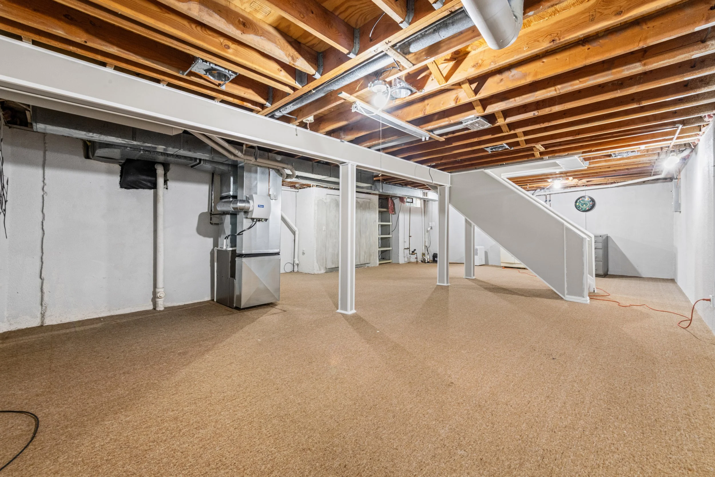 Empty basement with exposed wooden ceiling beams, white walls, furnace, ductwork, and beige carpeted floor.