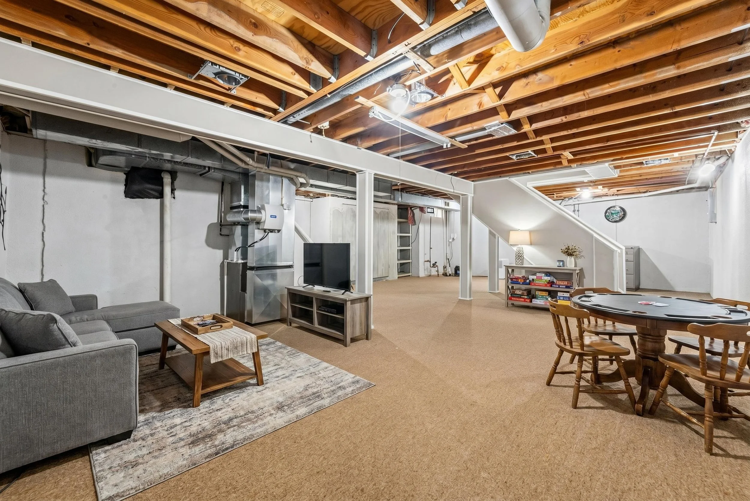 A large basement living area with a high wooden ceiling and various furniture including a gray sofa with pillows, a wooden coffee table, a TV stand with a small TV, a poker table, and a bookshelf with games. Exposed ductwork and pipes are visible alo