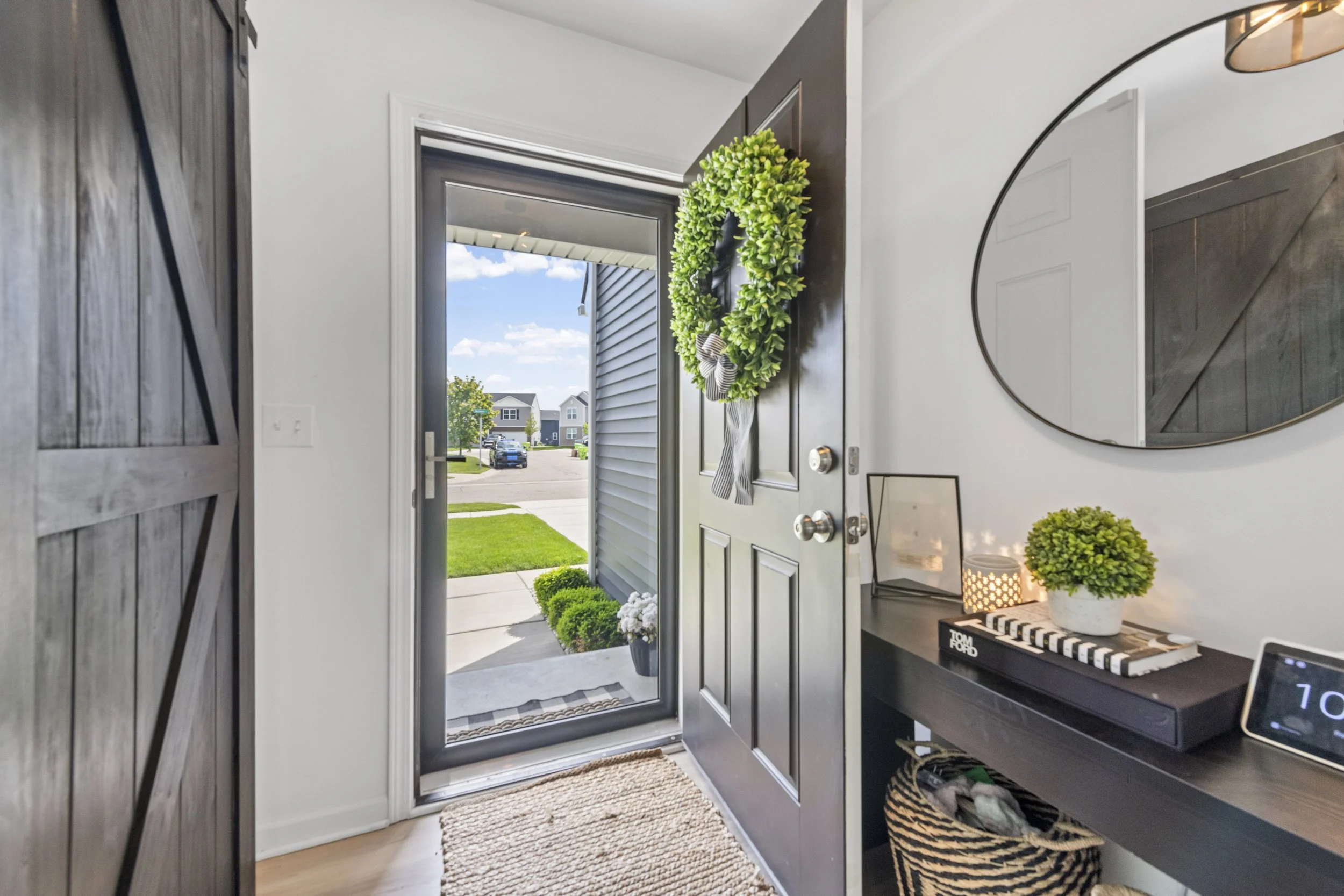 Interior view of a modern entryway with a door open to the outside, showing a street with houses and cars. The door is decorated with a green wreath with a bow. Inside, there is a small table with a potted plant, a candle, and a decorative item. A mi