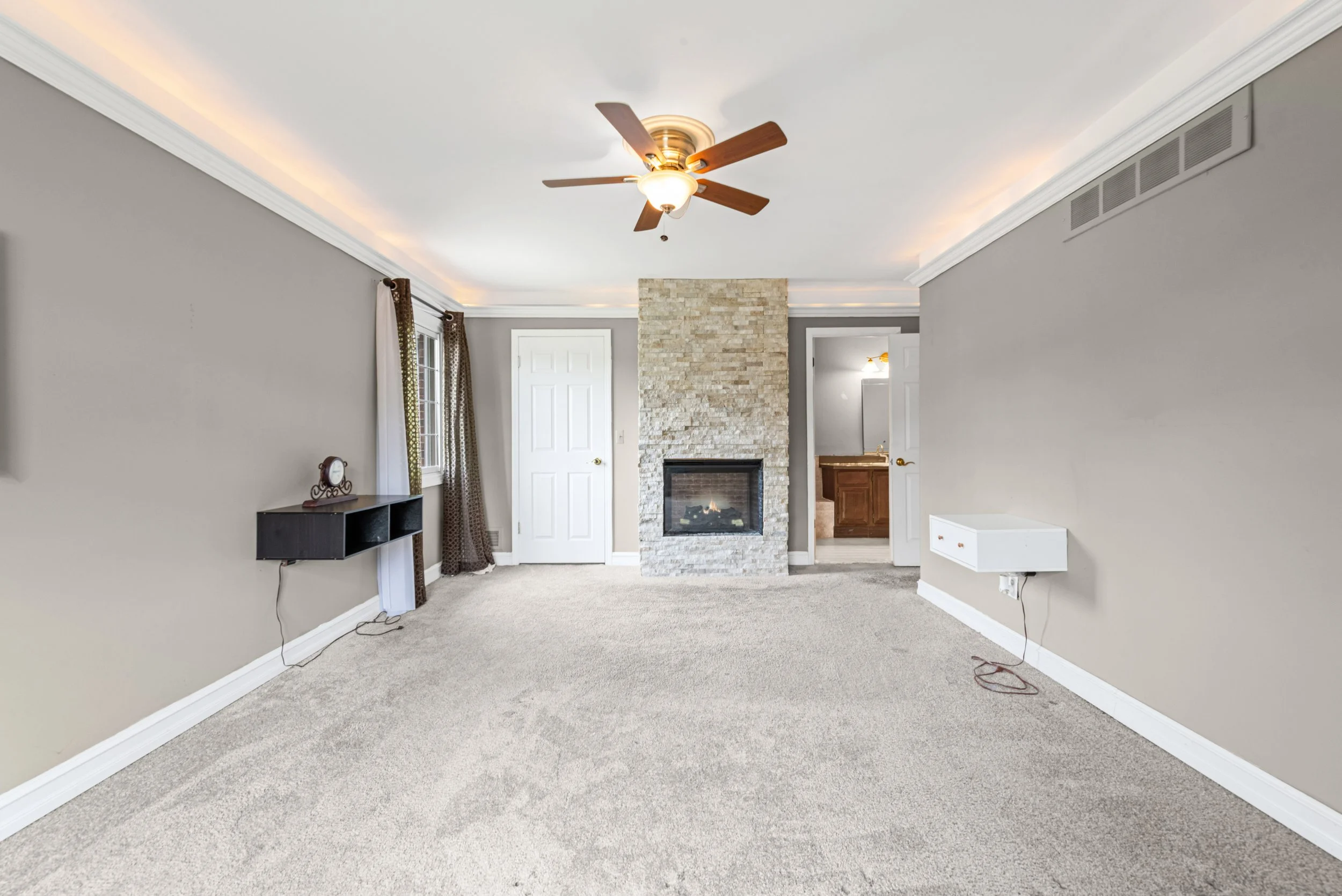 Empty living room with beige carpet, gray walls, a ceiling fan, a stone fireplace, white doors, and some curtains.