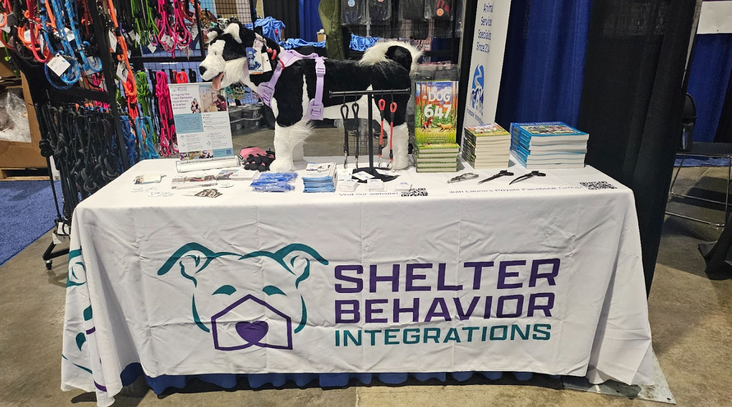 A Shelter Behavior Integrations table is set up at a conference. it's covered with animal handling tools, educational books, and a stuffed dog wearing a harness.