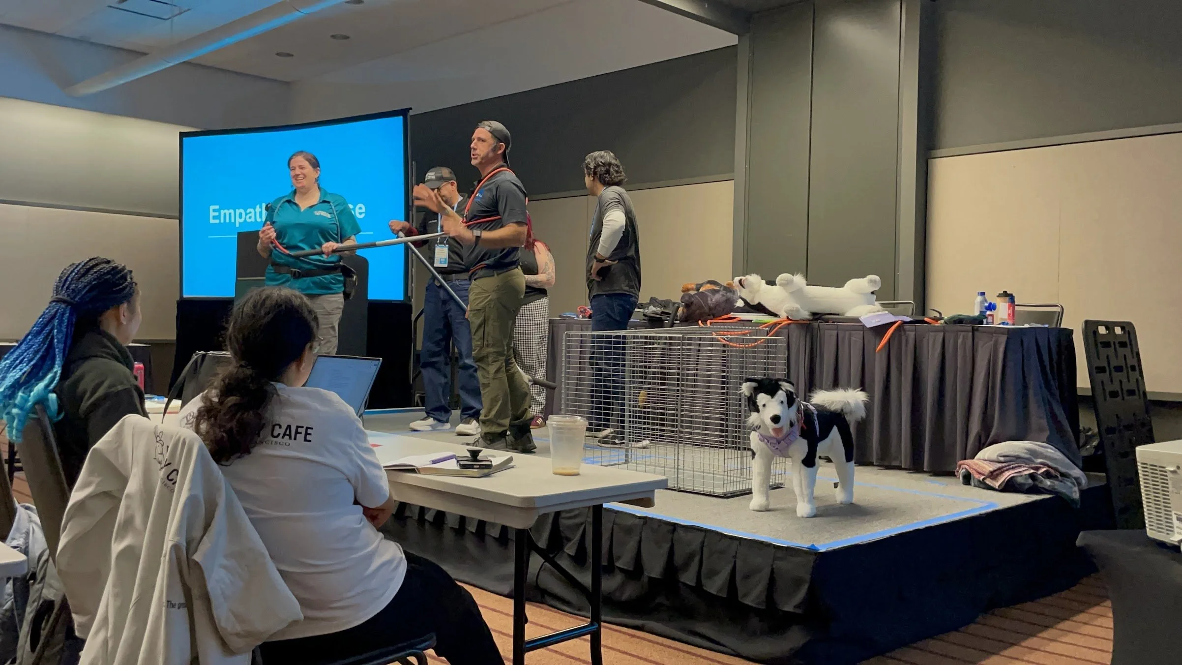 Laurie Lawless, John Peavler, and other animal care experts present an "empathy exercise" for an audience of animal welfare professionals. On stage, there is a large cage, a stuffed dog, and the named experts holding safety equipment.