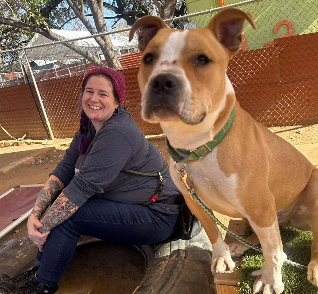 A brown-and-white dog sits on a wooden perch covered in fake grass beside Laurie, who sits and smiles on a tire. The pair sits outside in a play yard.