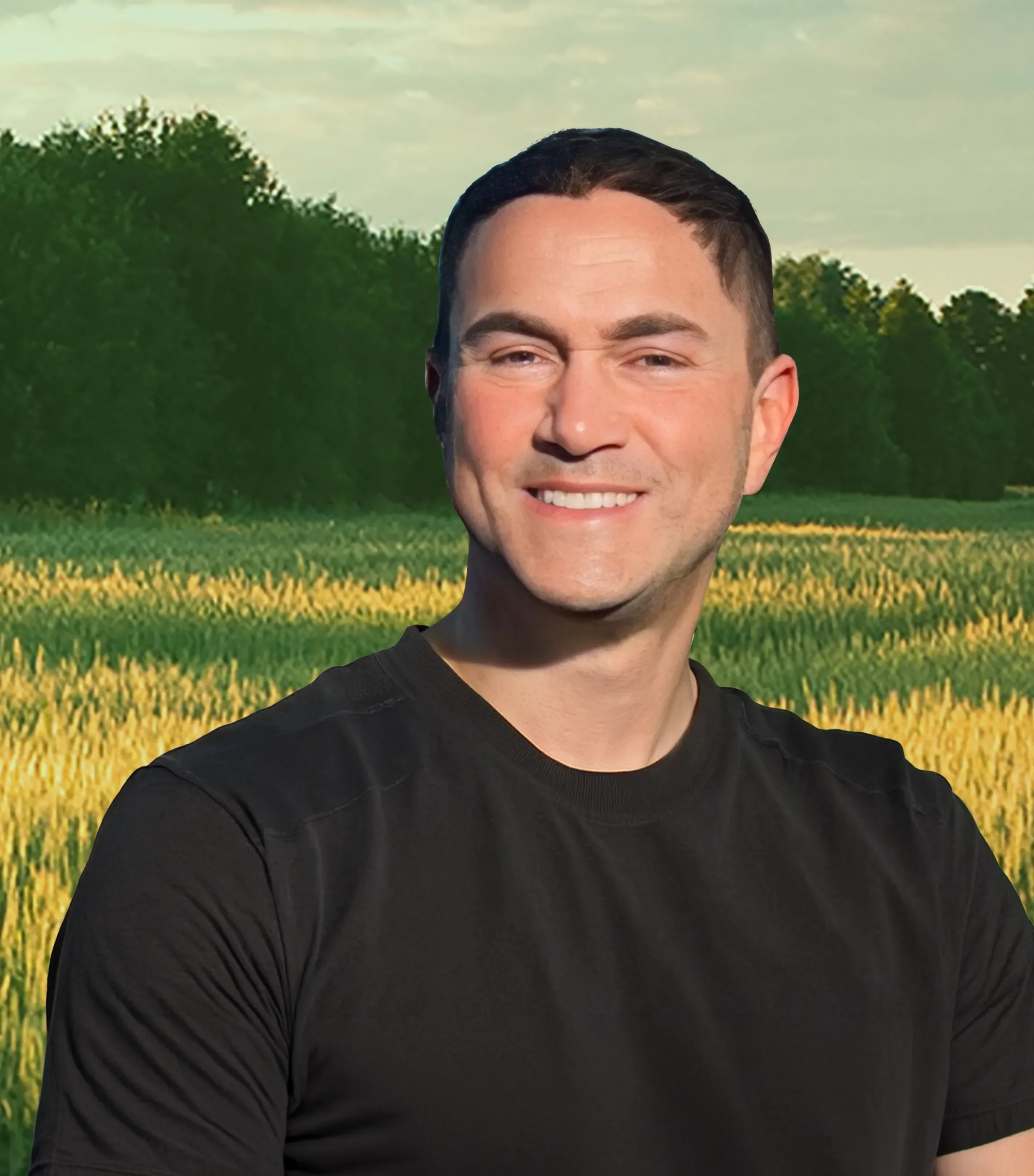 Close-up of a smiling young man with dark hair, wearing a black shirt, standing in front of a background of a green field with yellow flowers and trees, during daytime.