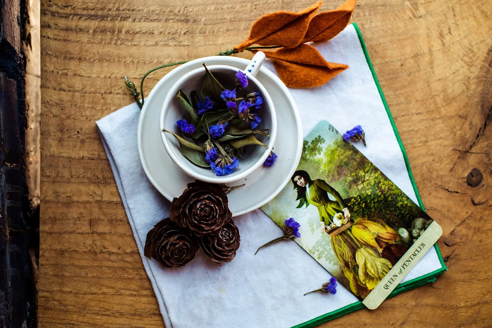 A white cup and saucer with dried purple flowers and green leaves, three brown pinecones, a folded tarot card titled "Queen of Pentacles" with illustration of a woman in a green outfit, on a wooden surface with a white cloth and dried orange leaves.