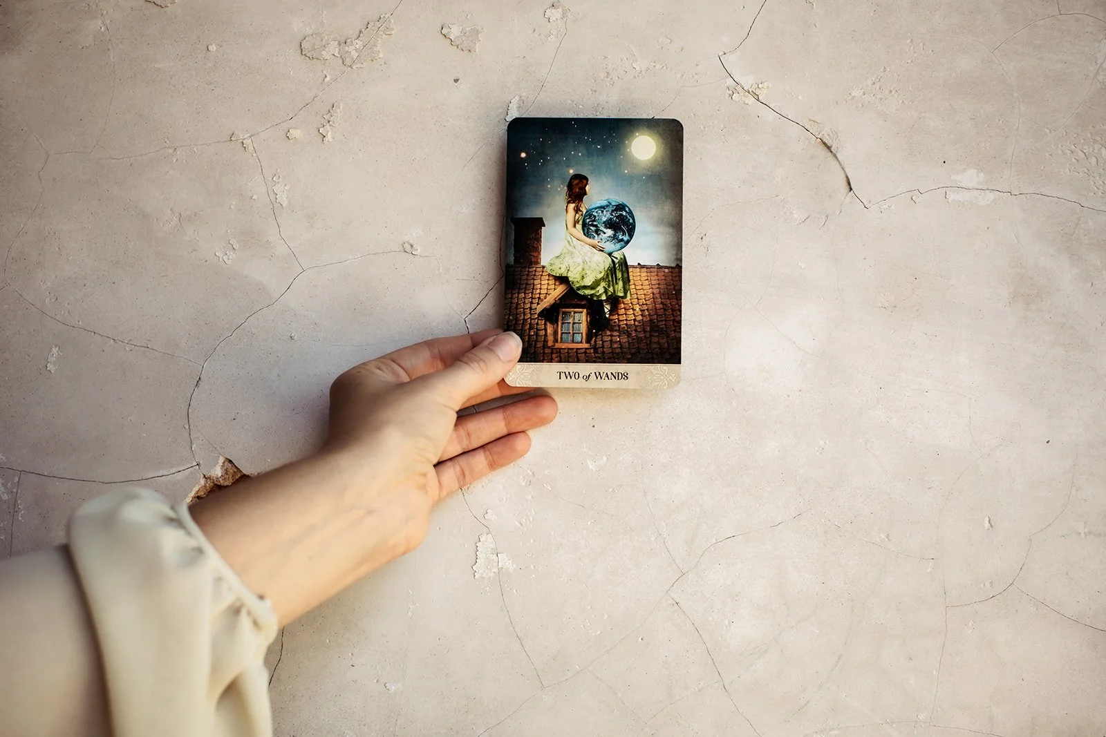 A tarot card titled "Two of Wands" being held over a cracked, beige surface. The card depicts a woman sitting on a rooftop at night, holding a globe, with a moon and stars in the sky.
