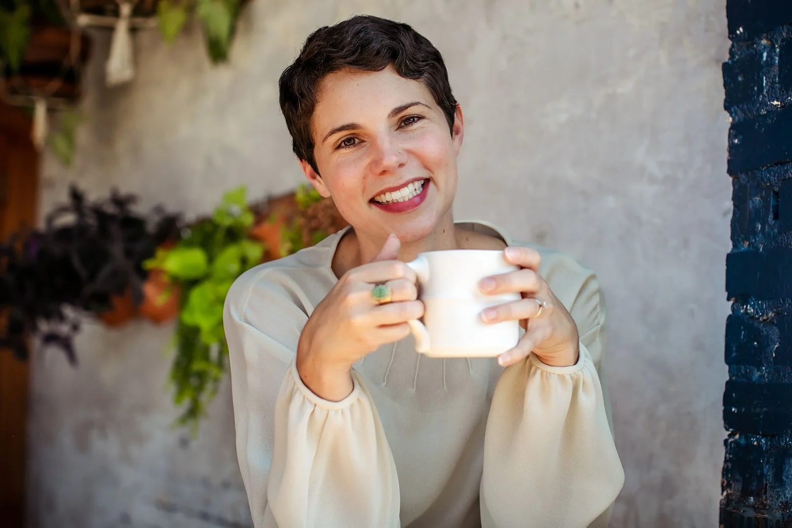 Woman with short dark hair smiling and holding a white mug outside against a textured background.