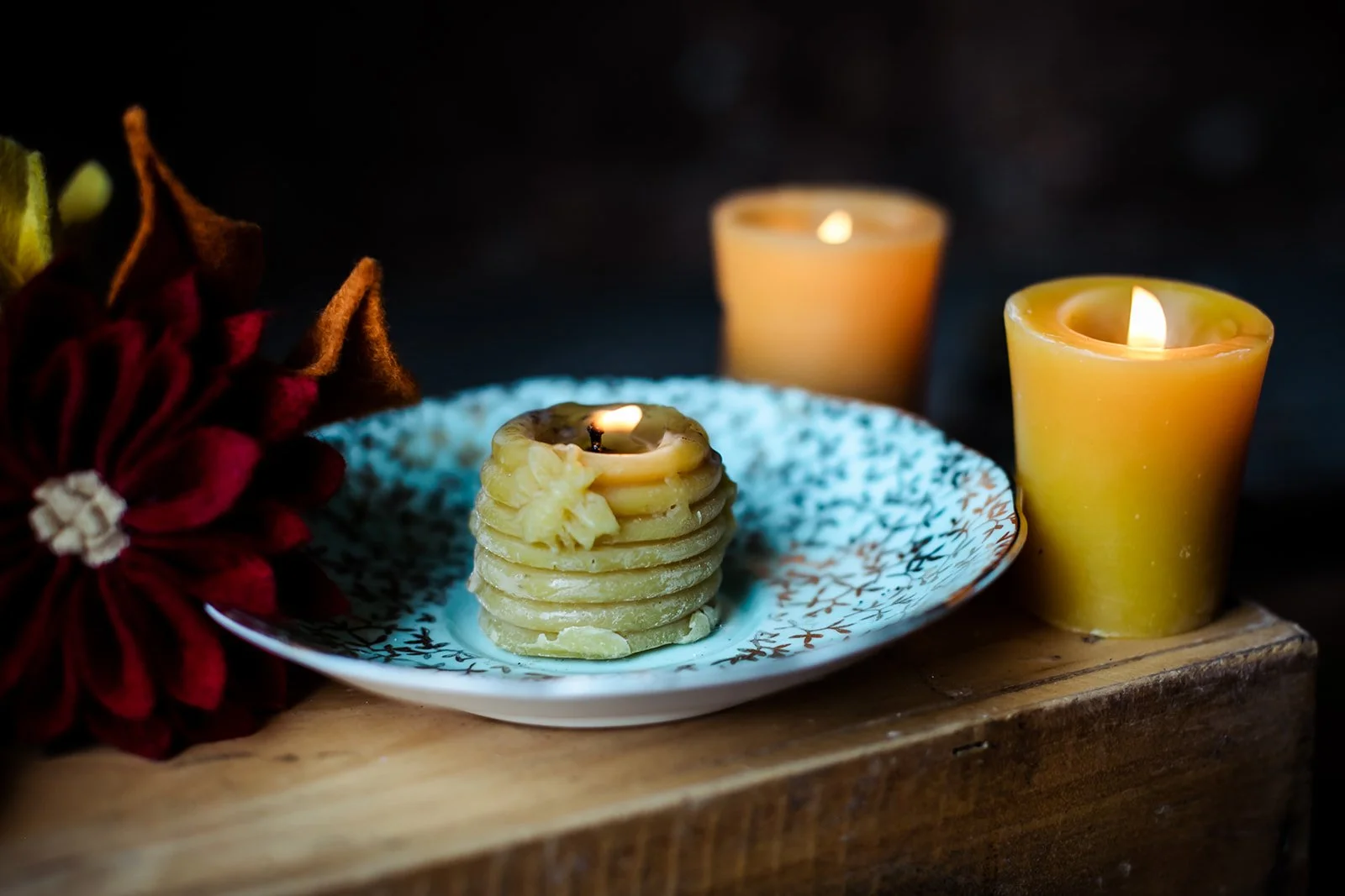A holiday scene with a plate of stacked wax melts shaped like a pine cone, two lit candles, and a red felt flower decoration on a wooden surface.