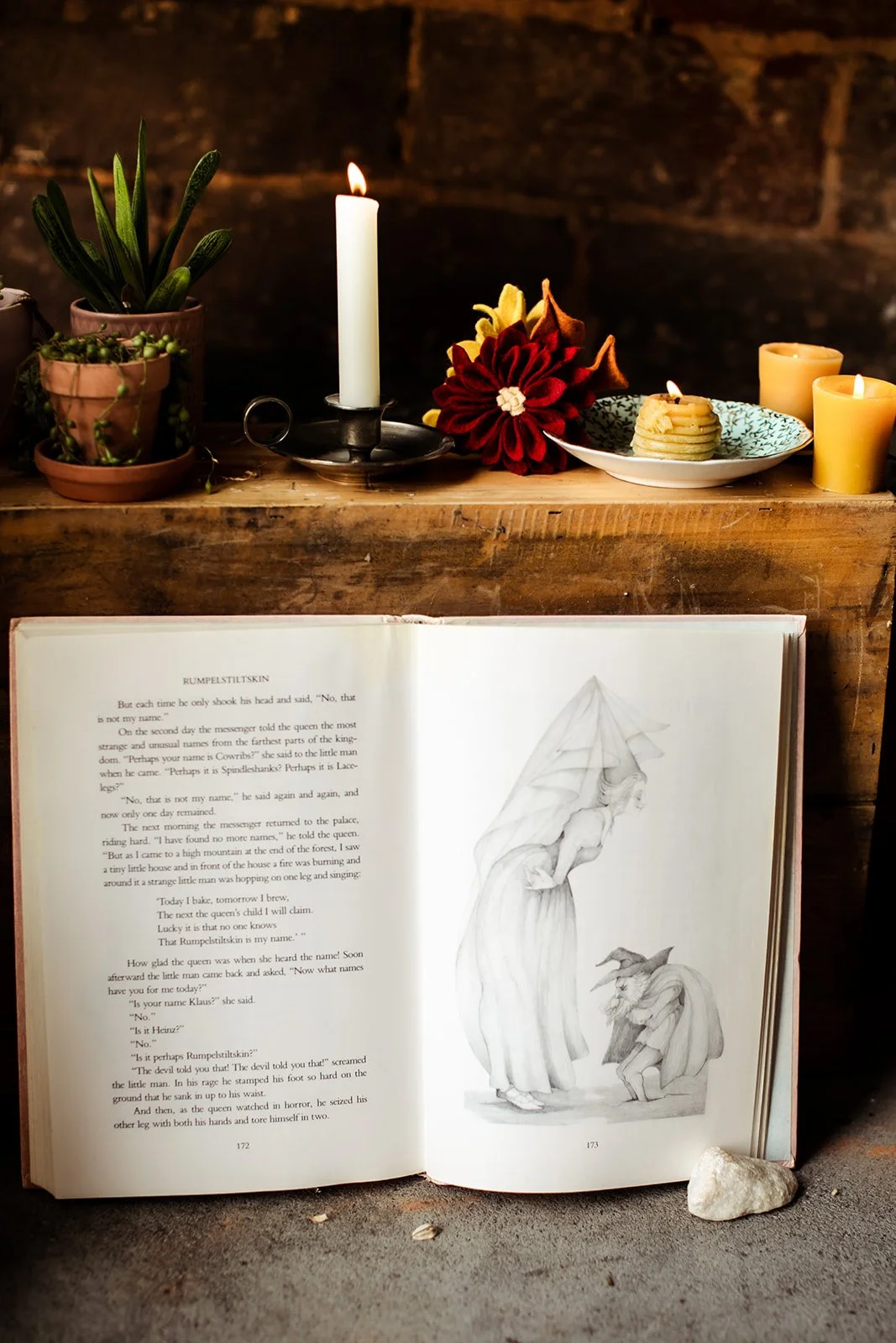 Open book with a sketched illustration of a woman in a flowing dress and a humanoid creature with horns, placed on a rustic surface; above it are candles, a potted plant, flowers, and a stone.