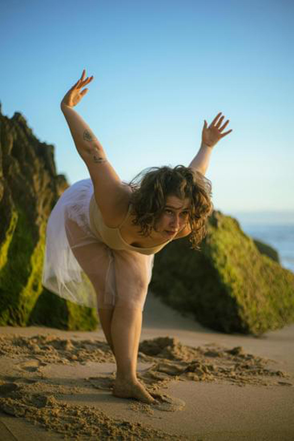 Woman performing a yoga pose on the beach during sunset with rocks and ocean in the background.