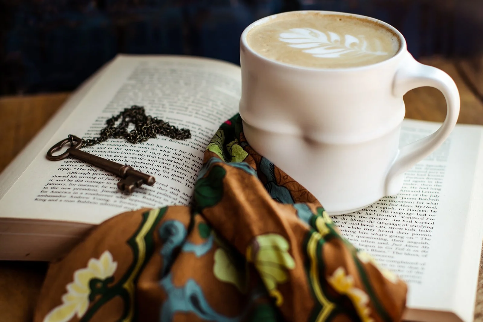 A white ceramic mug with latte art on a open book, a necklace with a key pendant, a colorful scarf in front, all on a wooden table.