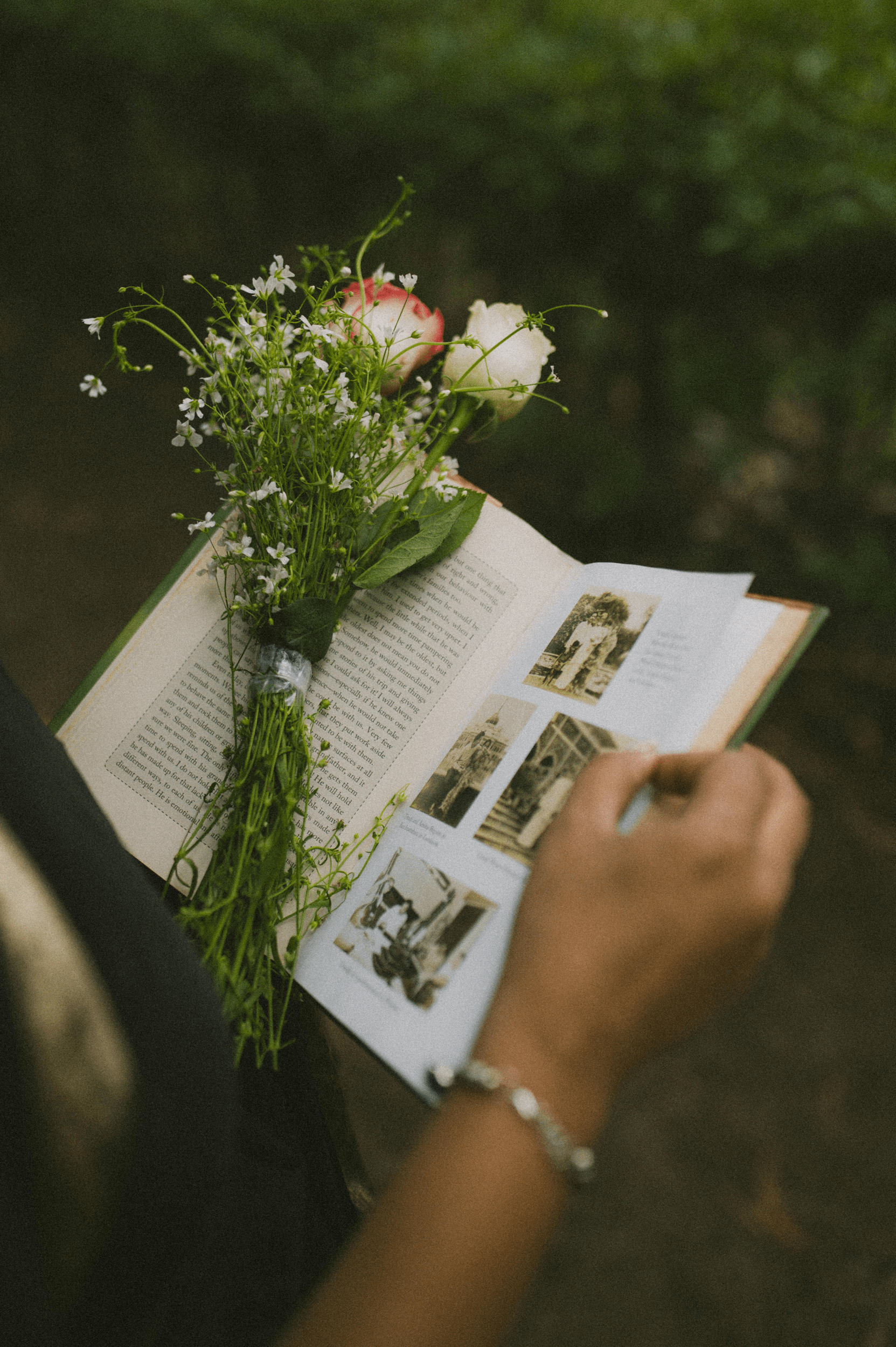 A person with a bracelet on their wrist is reading a book outdoors, with a bouquet of flowers resting on the open pages.