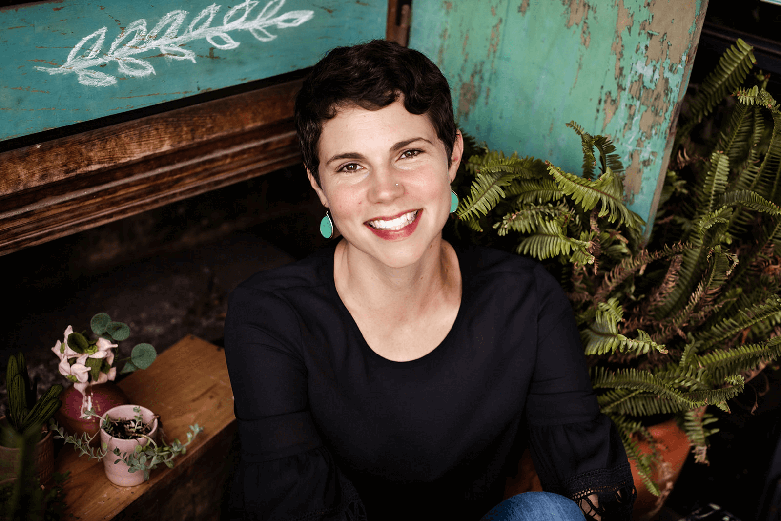 A woman with short dark hair, wearing a black top and turquoise earrings, is smiling while sitting among potted plants and outdoor decor.