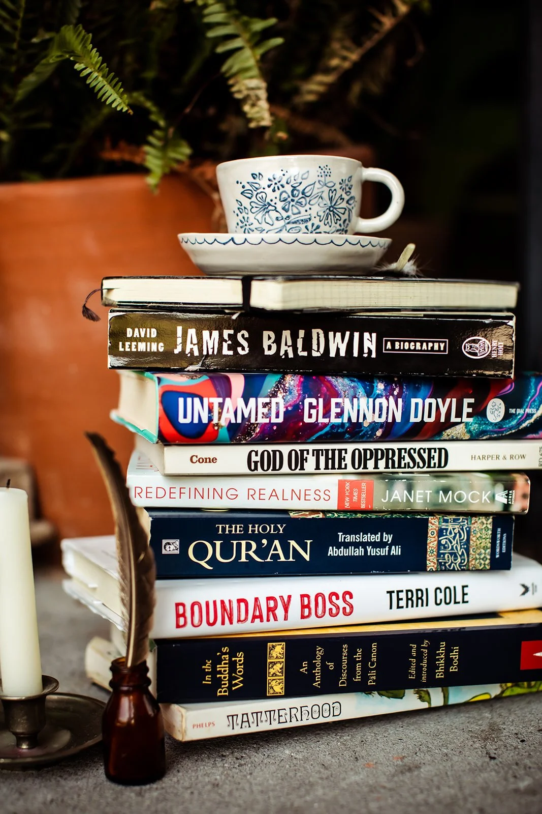 Stack of books topped with a ceramic cup and saucer, near a candle and a small vase with a feather, in front of a large plant.