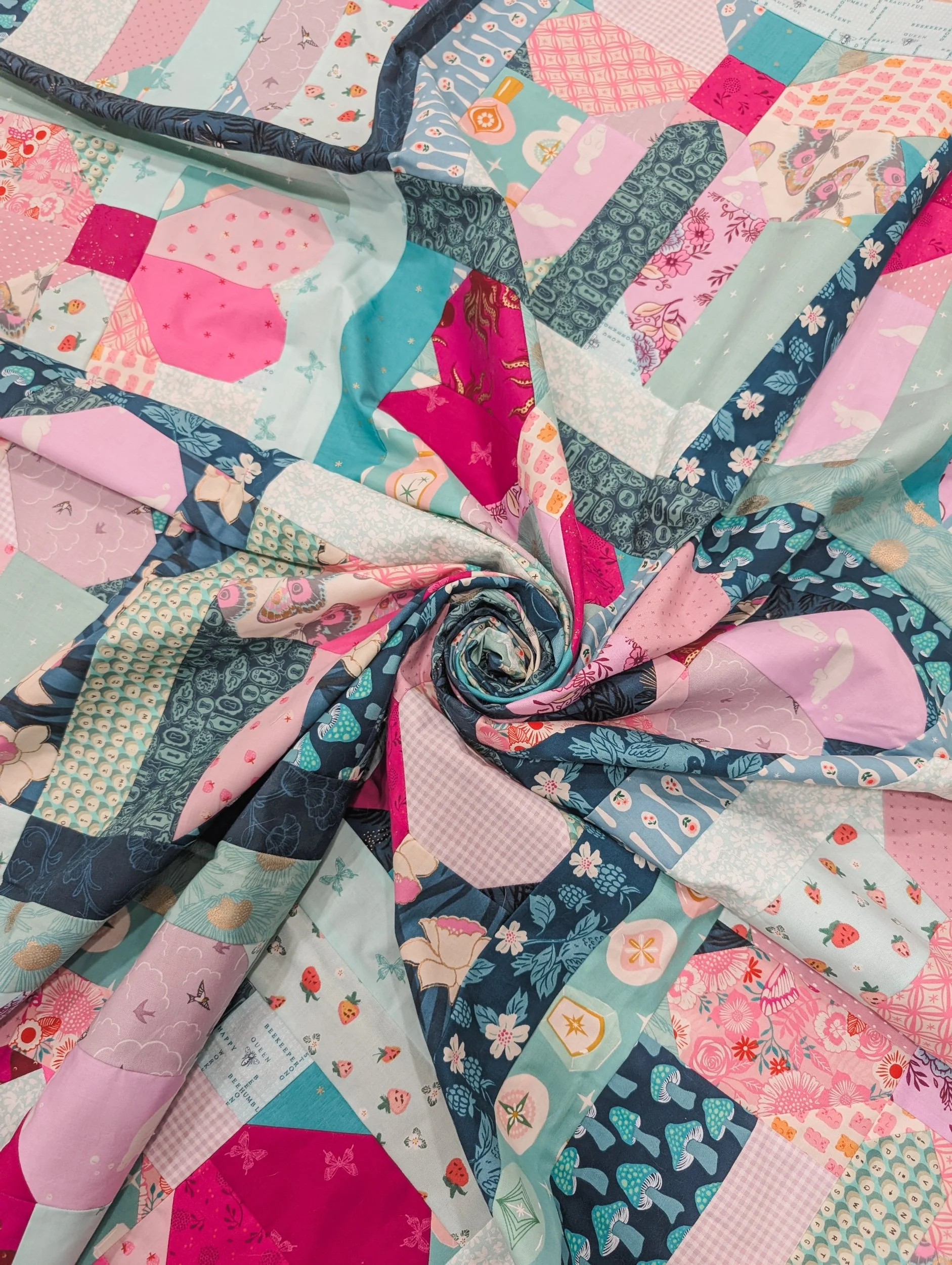 Close-up of a colorful quilt with various patterned fabric patches in pink, blue, and white, arranged in a spiral with some fabric swirling around.