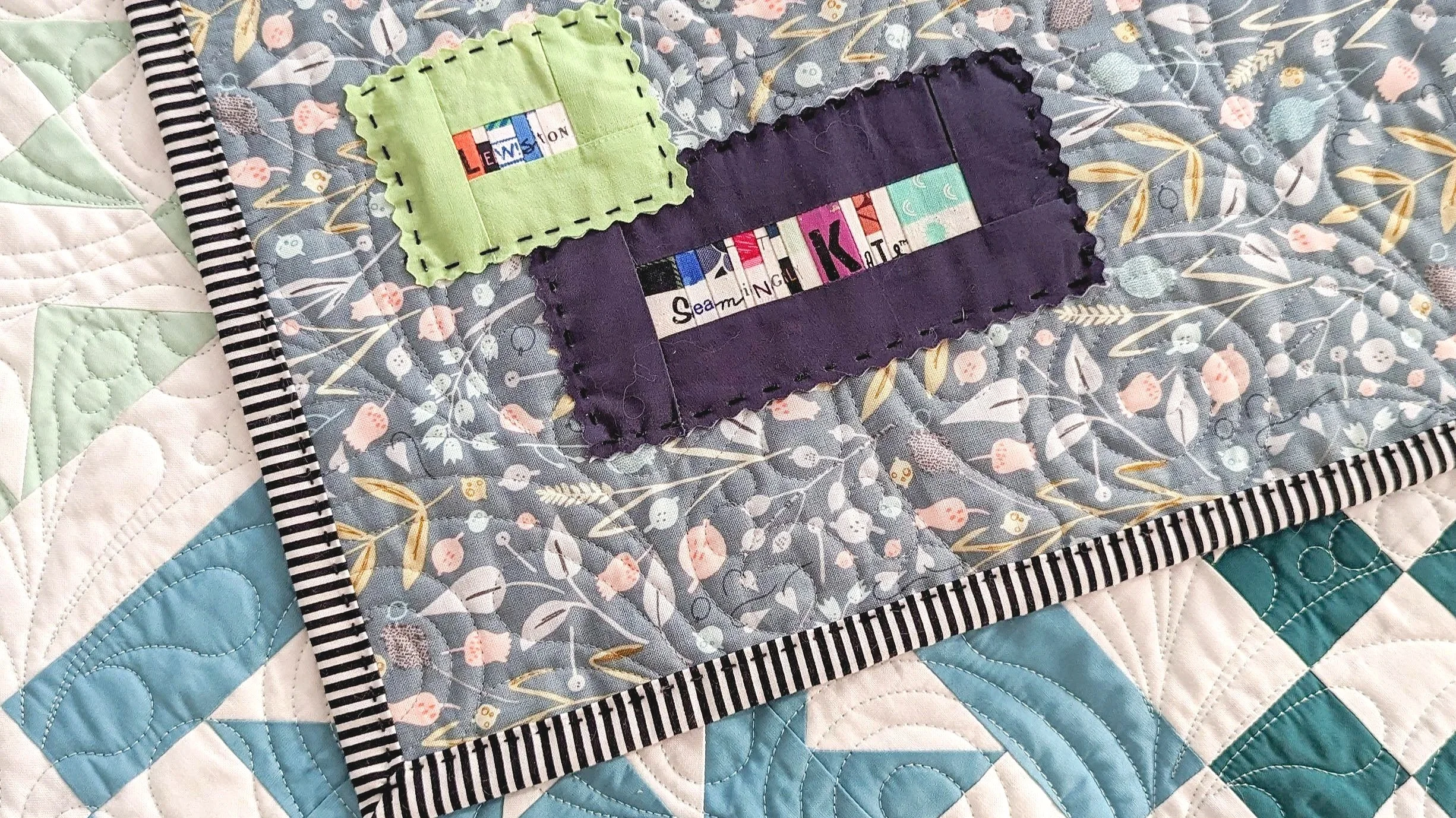 Close-up of a quilt with a floral pattern quilted in blue, white, and teal colors. Two fabric tags, one green and one purple, are sewn onto the quilt, with the purple tag containing the words 'Sewing K' and other partial text. The quilt has a black and white striped border.