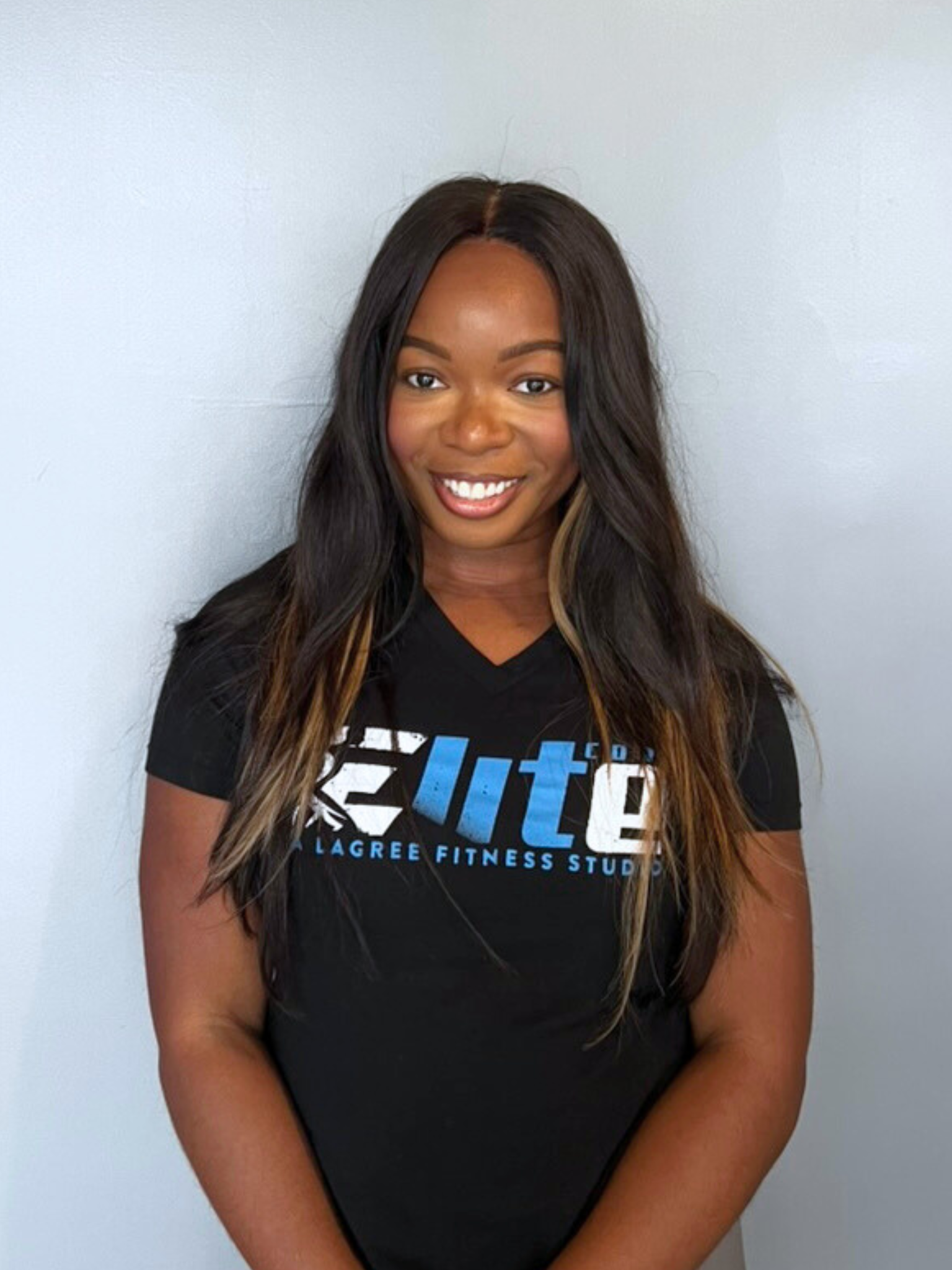 A young woman with long dark hair and light highlights, smiling, wearing a black T-shirt with blue and white text that reads 'Elite' and 'Lagree Fitness Studio', standing against a plain light-colored wall.