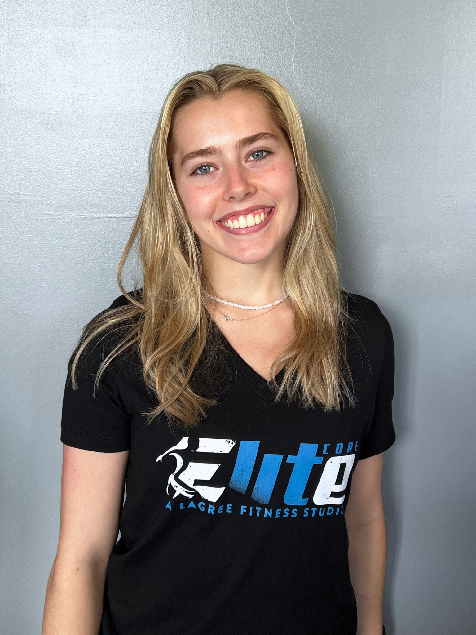A young woman with blonde hair, wearing a black t-shirt with blue and white text that says 'Lite Core' and 'A Lagree Fitness Studio', smiling and standing against a plain light grey wall.