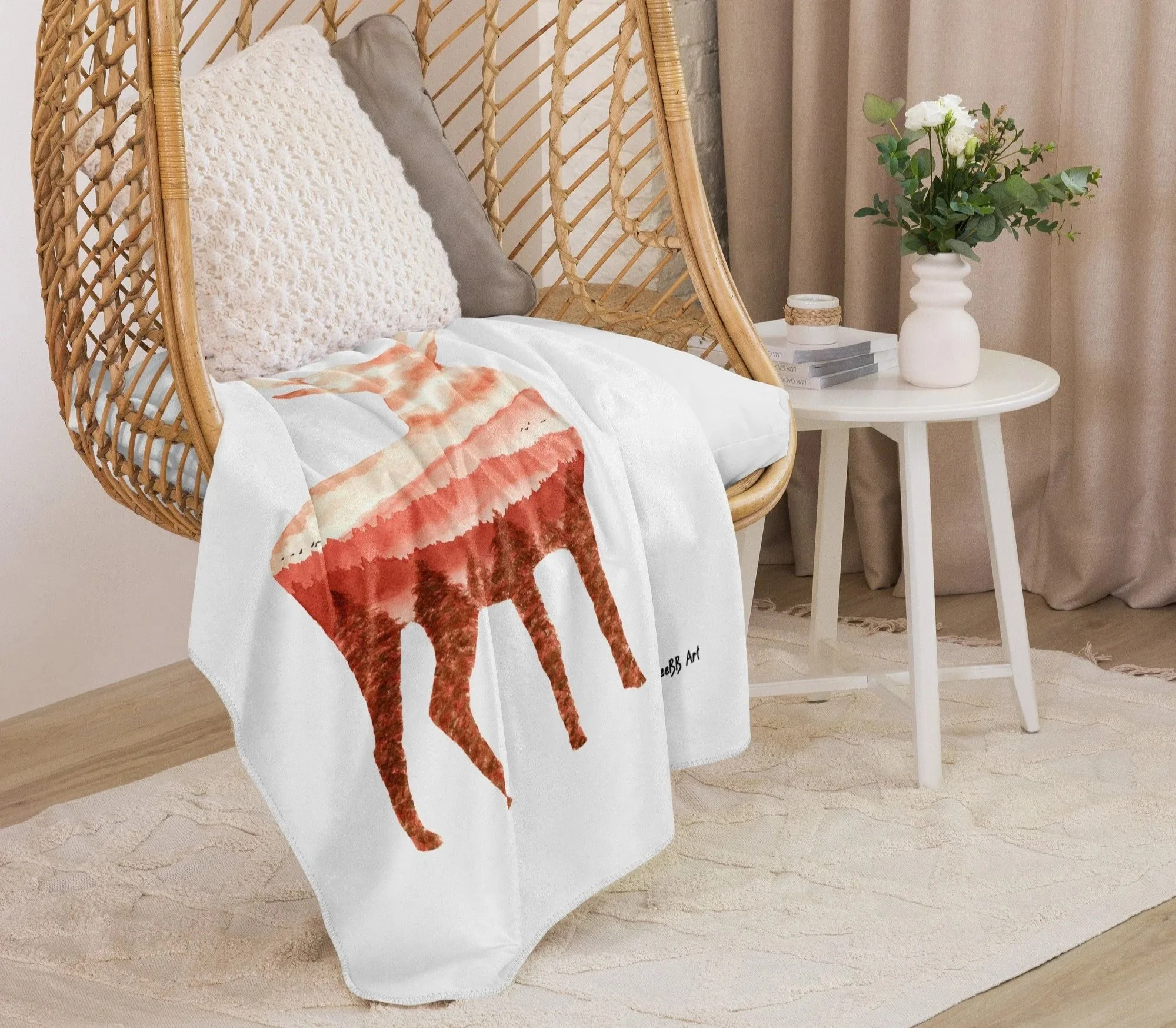 Close-up of a wicker hanging chair with cushions, a white blanket with a red stag print, beside a side table with a stack of books, a white vase with green leaves and white flowers, set in a room with beige curtains and a light-colored rug.
