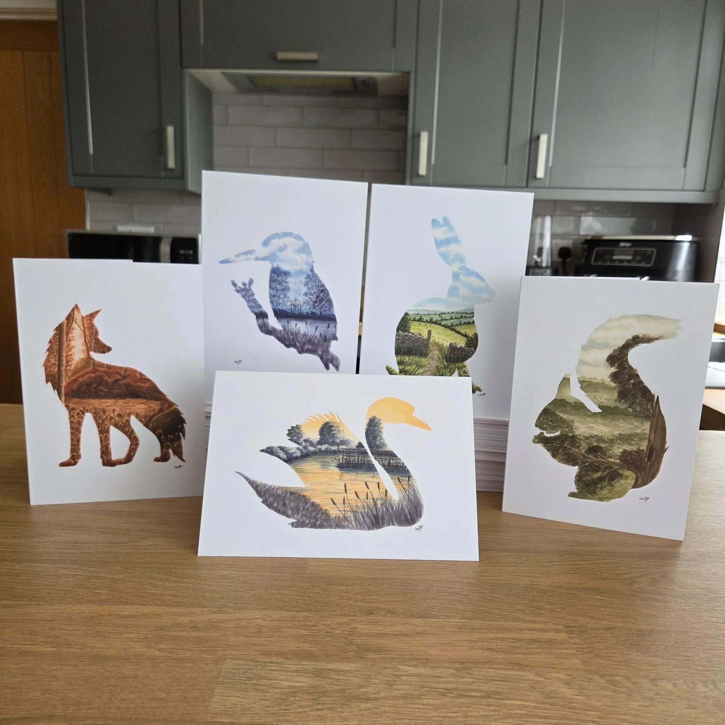 A collection of 5 greeting cards of animals in watercolour, including a bear, a kingfisher, and a fox, displayed on white papers on a wooden surface in a room with gray cabinets and stainless steel appliances.