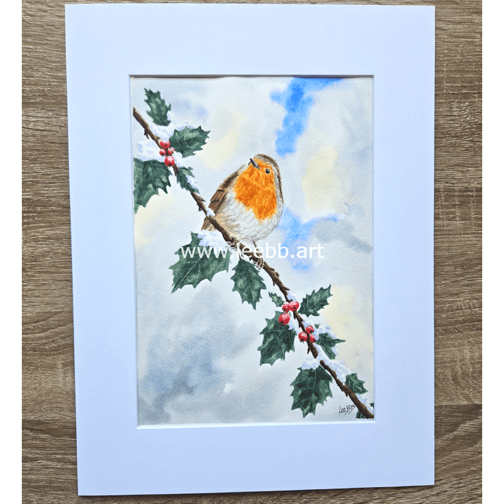 Watercolor painting of a robin perched on a holly branch with red berries, snowy background, inside a white mat frame on a wooden surface.