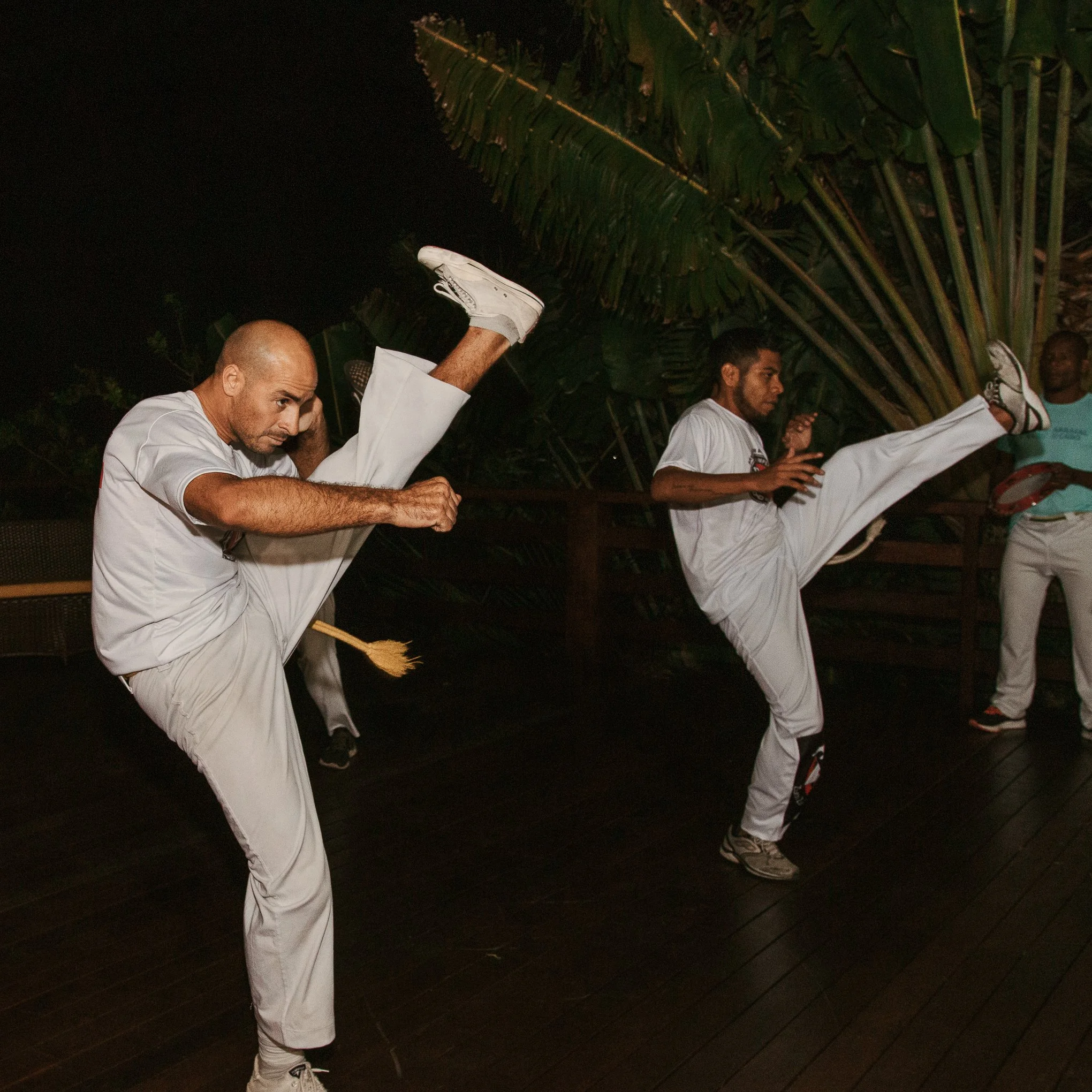 Two men practicing martial arts and executing high kicks indoors, accompanied by a person in the background holding a tambourine.